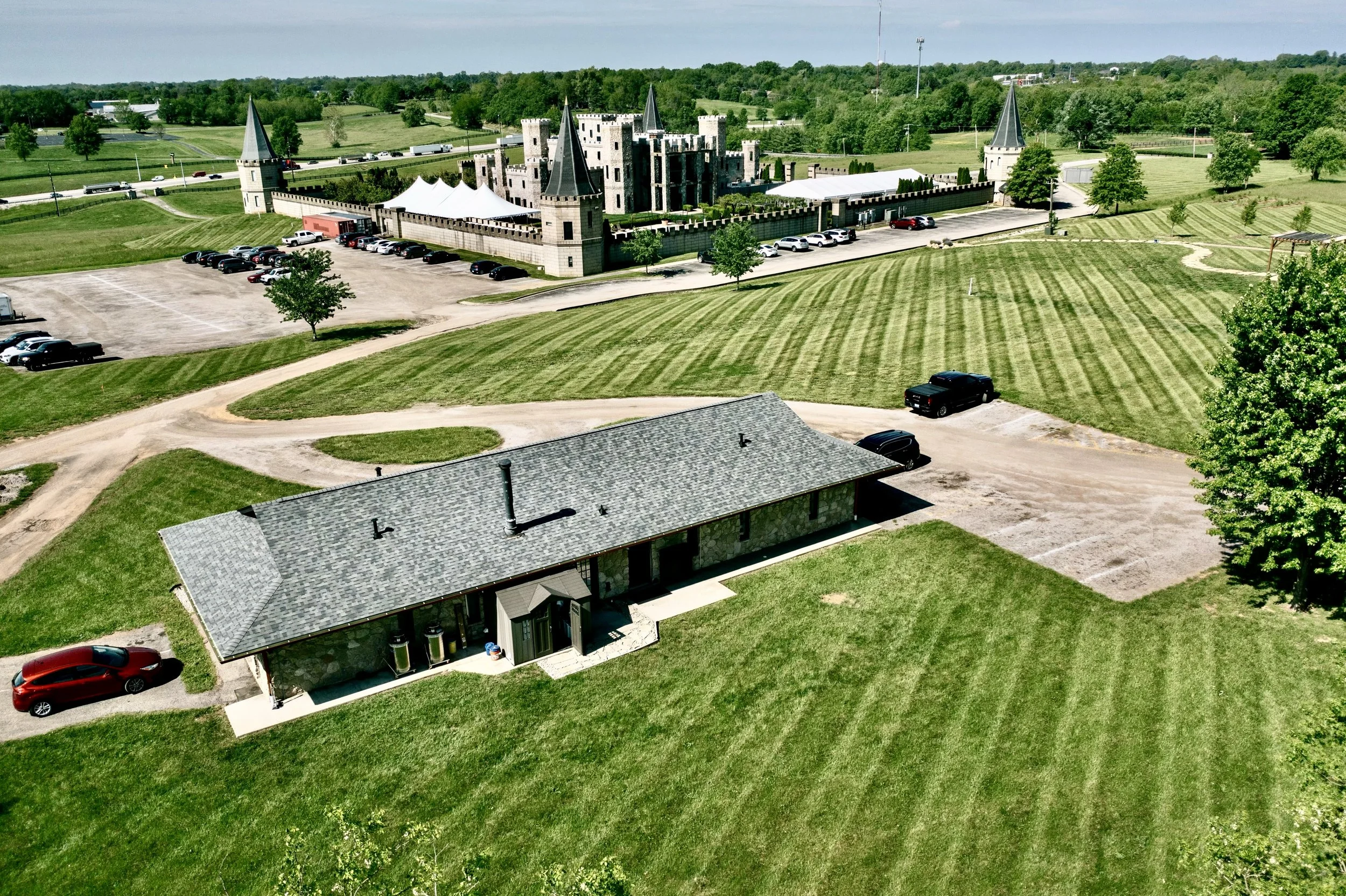 A castle with four turrets, surrounded by a wall, located behind a large field with striped grass, and a building with a gray roof in the foreground. Several cars are parked around the area, and there is a parking lot near the castle. The background 