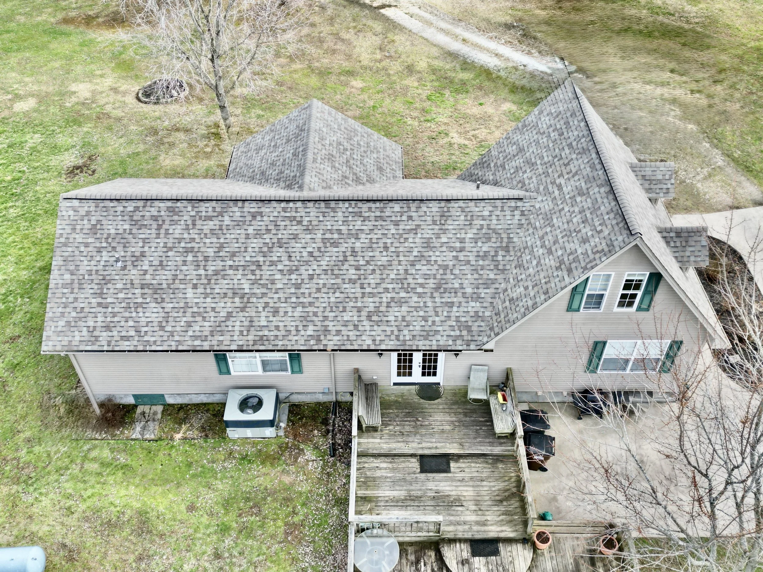 An aerial view of a house with a wooden deck, green shutters, and a shingled roof surrounded by grass, trees, and a small waterway.