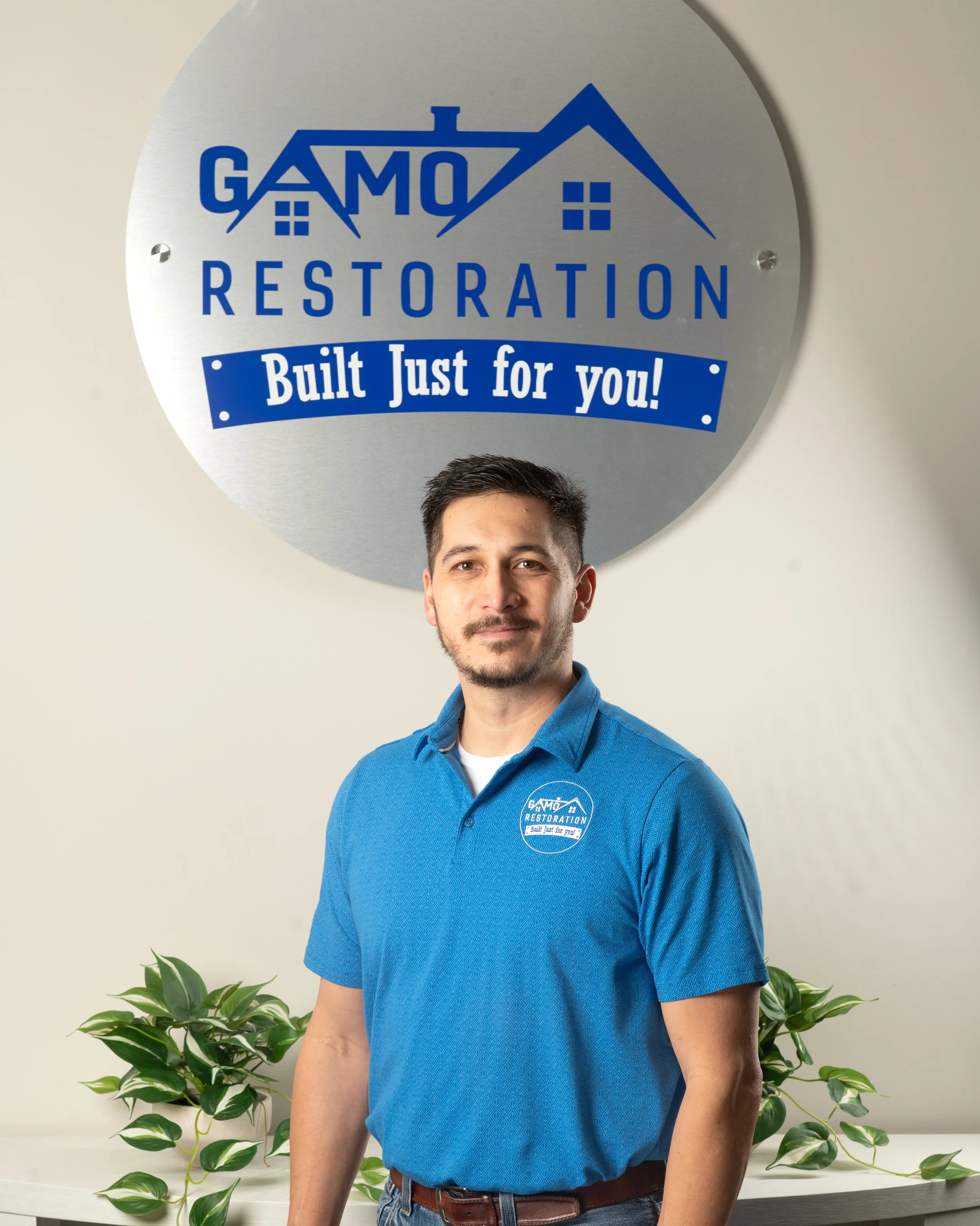 A man in a blue polo shirt standing in front of a wall with a large sign that reads 'GAMO Restoration Built Just for you!'. There are green plants on a white table behind him.