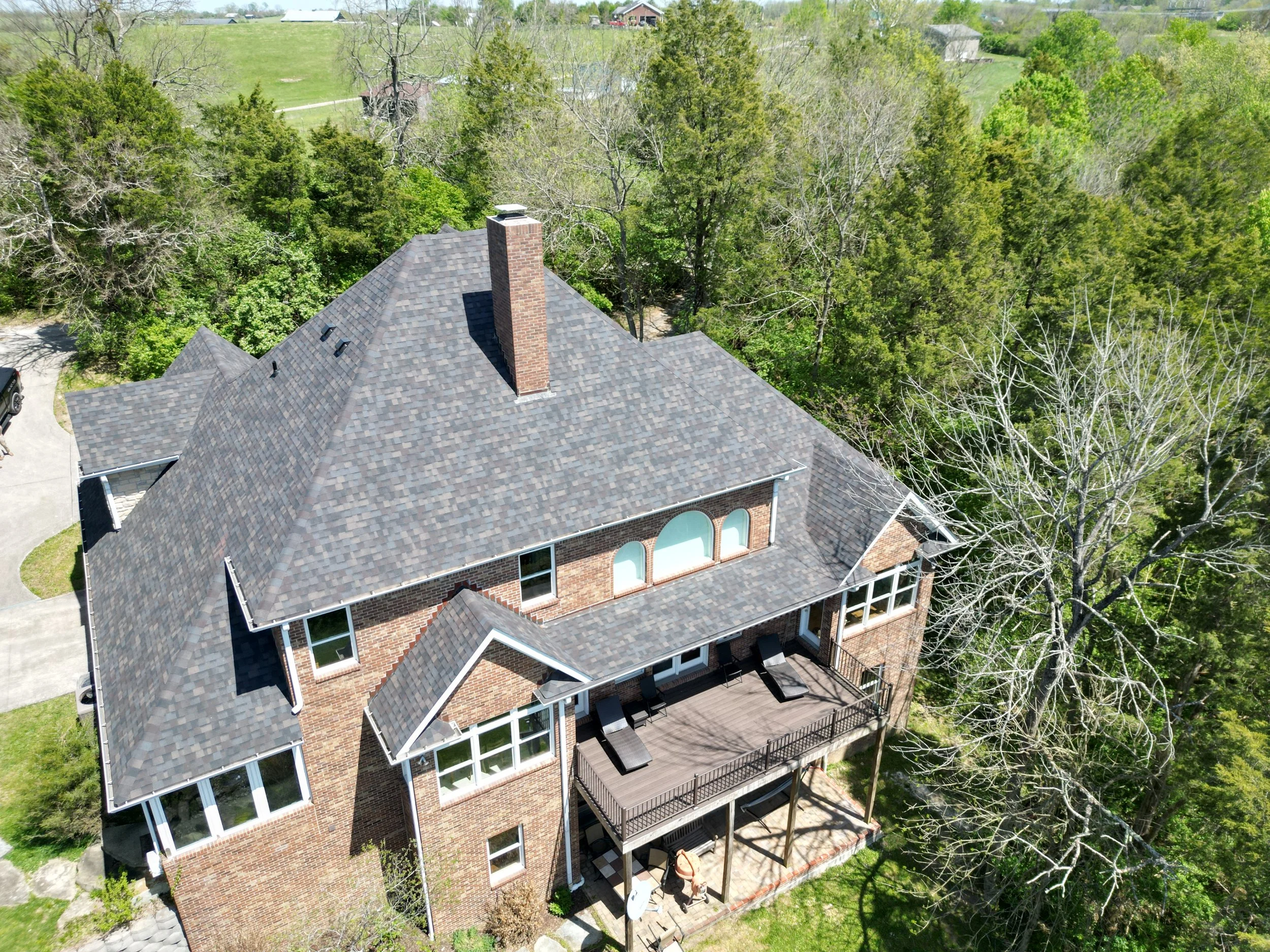 An aerial view of a two-story brick house with a large, multi-gabled shingle roof, chimney, and a metal balcony with outdoor furniture surrounded by trees and greenery.