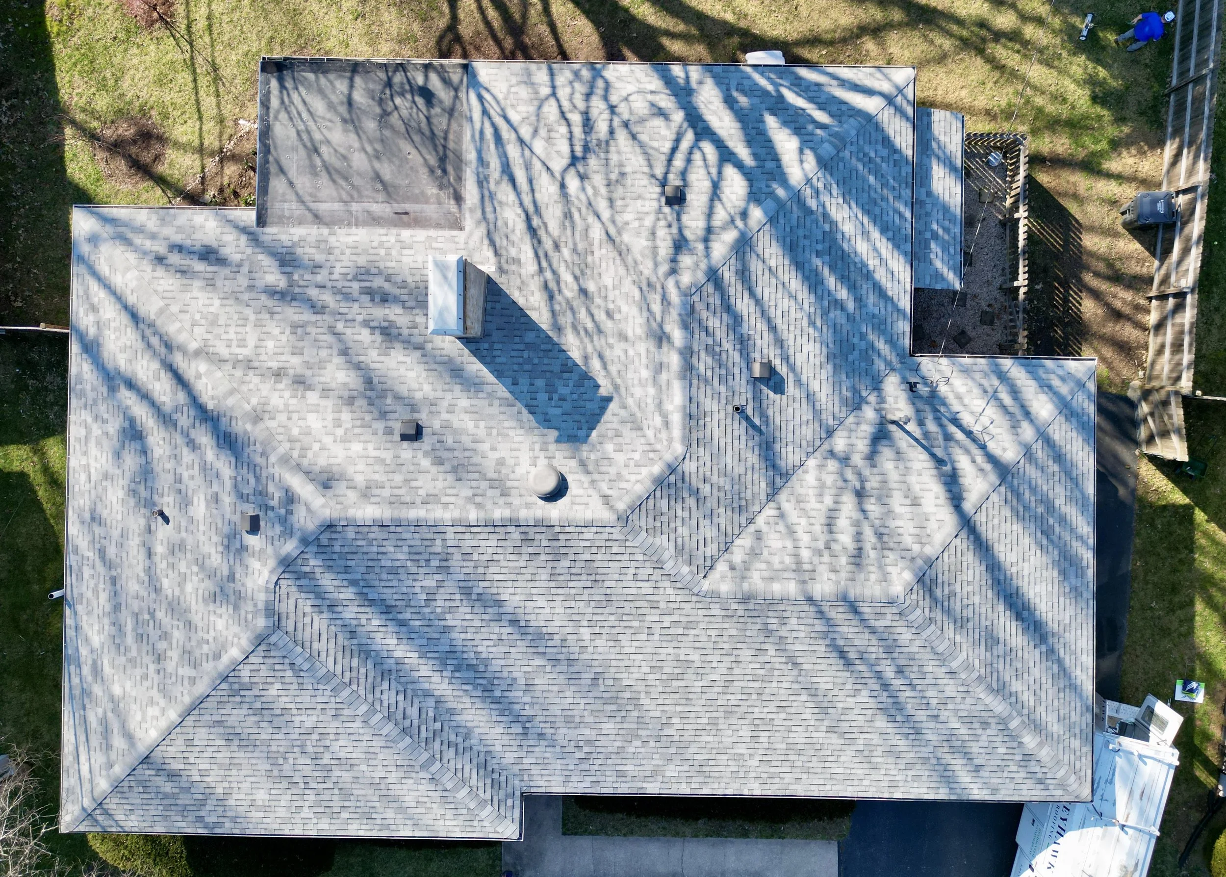 Aerial view of a house roof covered with light-colored shingles, surrounded by a grassy yard and some trees, with shadows cast by the trees on the roof.