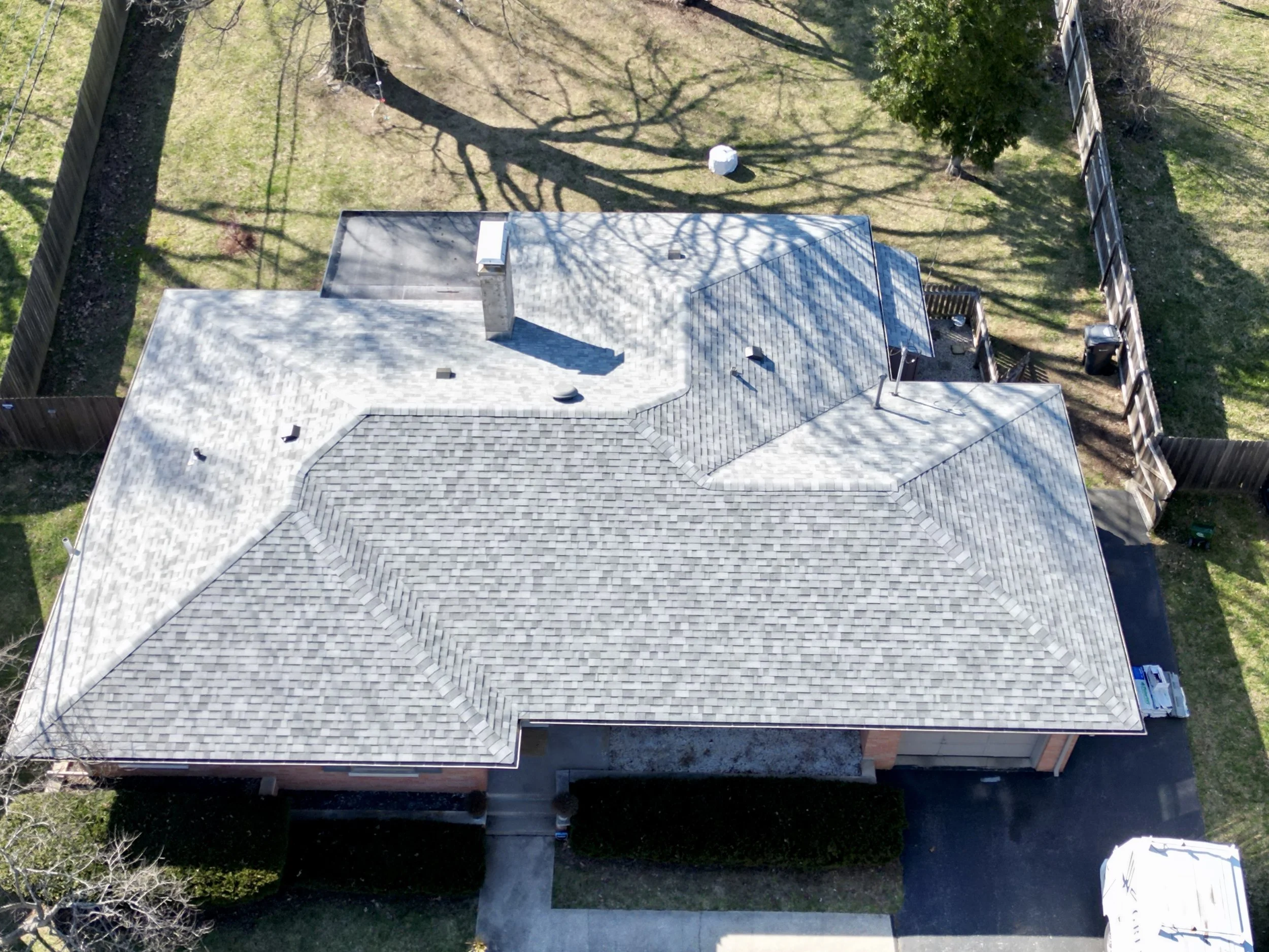 Aerial view of a house with a complex shingle roof surrounded by a backyard with trees, a fence, and a paved driveway.