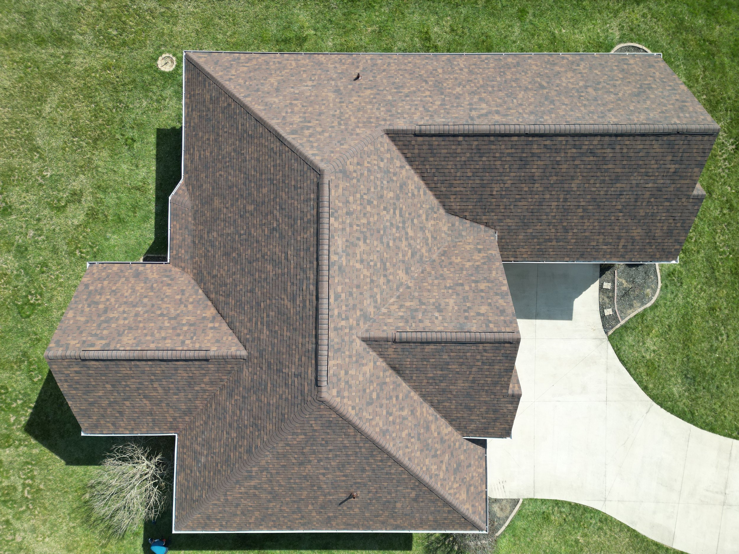 Aerial view of a house with brown shingle roof, surrounding green lawn, concrete driveway, and a small tree in the yard.