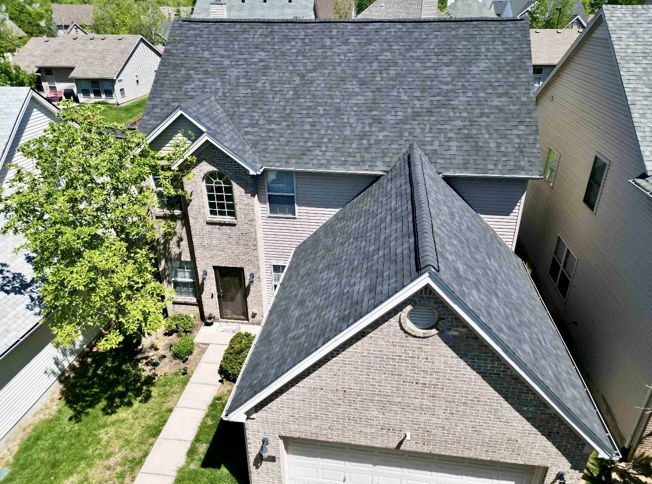 Aerial view of a residential house with brick and siding exterior, gray shingle roof, front yard with pathway, and neighboring houses.