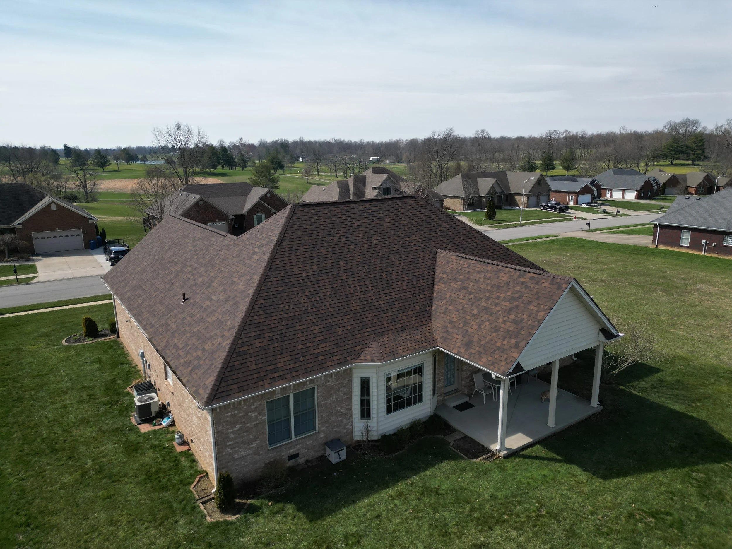 An aerial view of a suburban neighborhood with multiple houses, including a brick house with a covered patio in the foreground, and a golf course or park in the background under a cloudy sky.