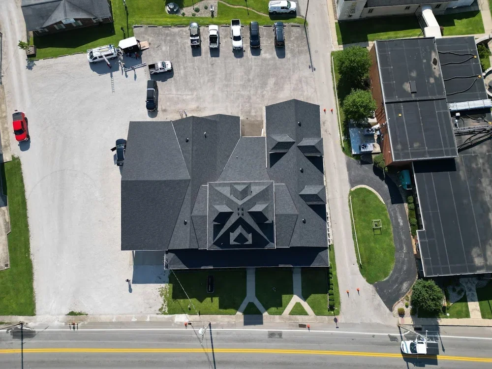 Aerial view of a building with a dark gray roof, surrounded by parking lots, grassy areas, and neighboring buildings. There are several cars parked and a few on the street.