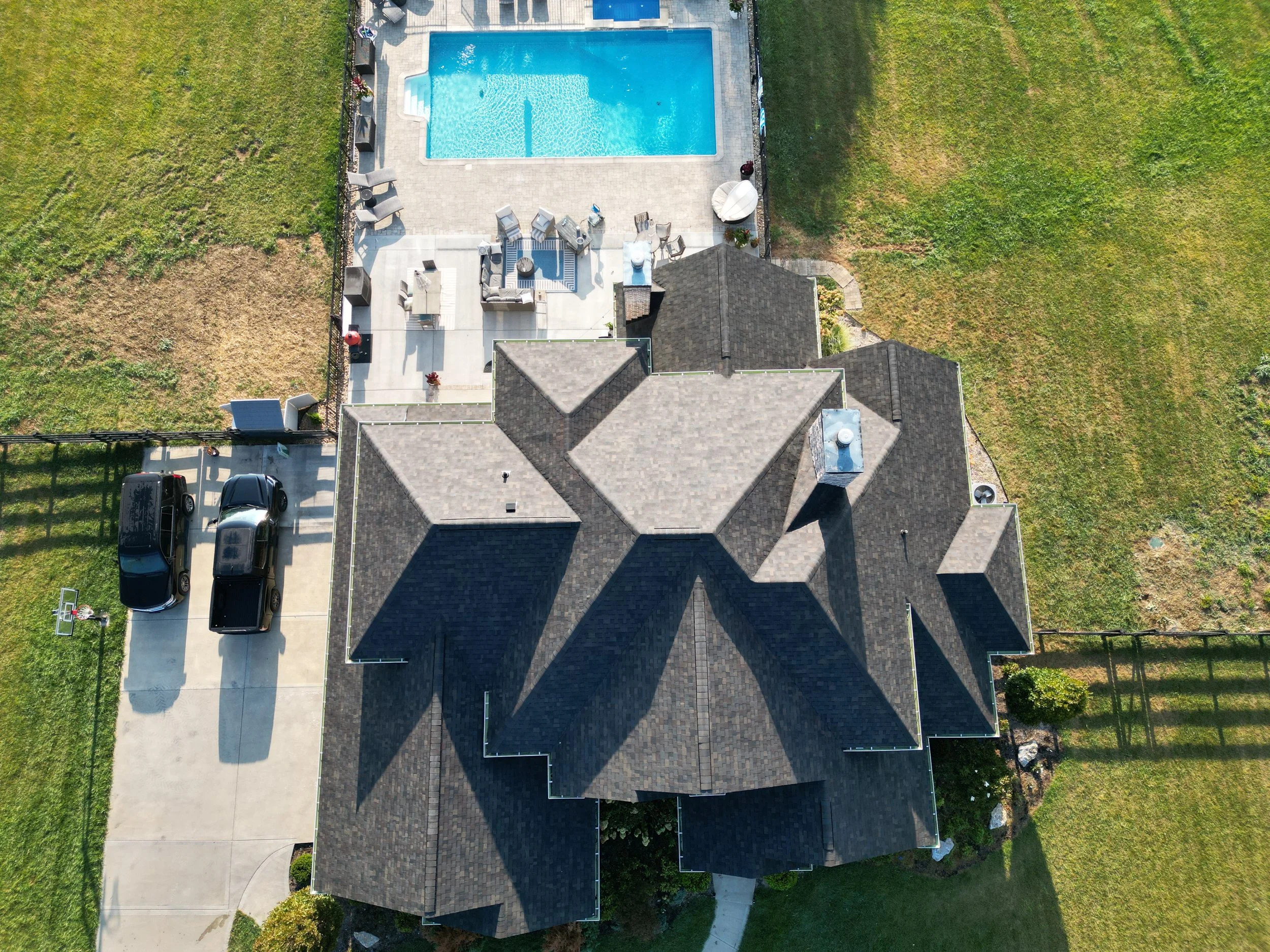 An aerial view of a house with a multi-gabled roof, a backyard with a swimming pool, outdoor seating, and a driveway with two cars parked side by side.