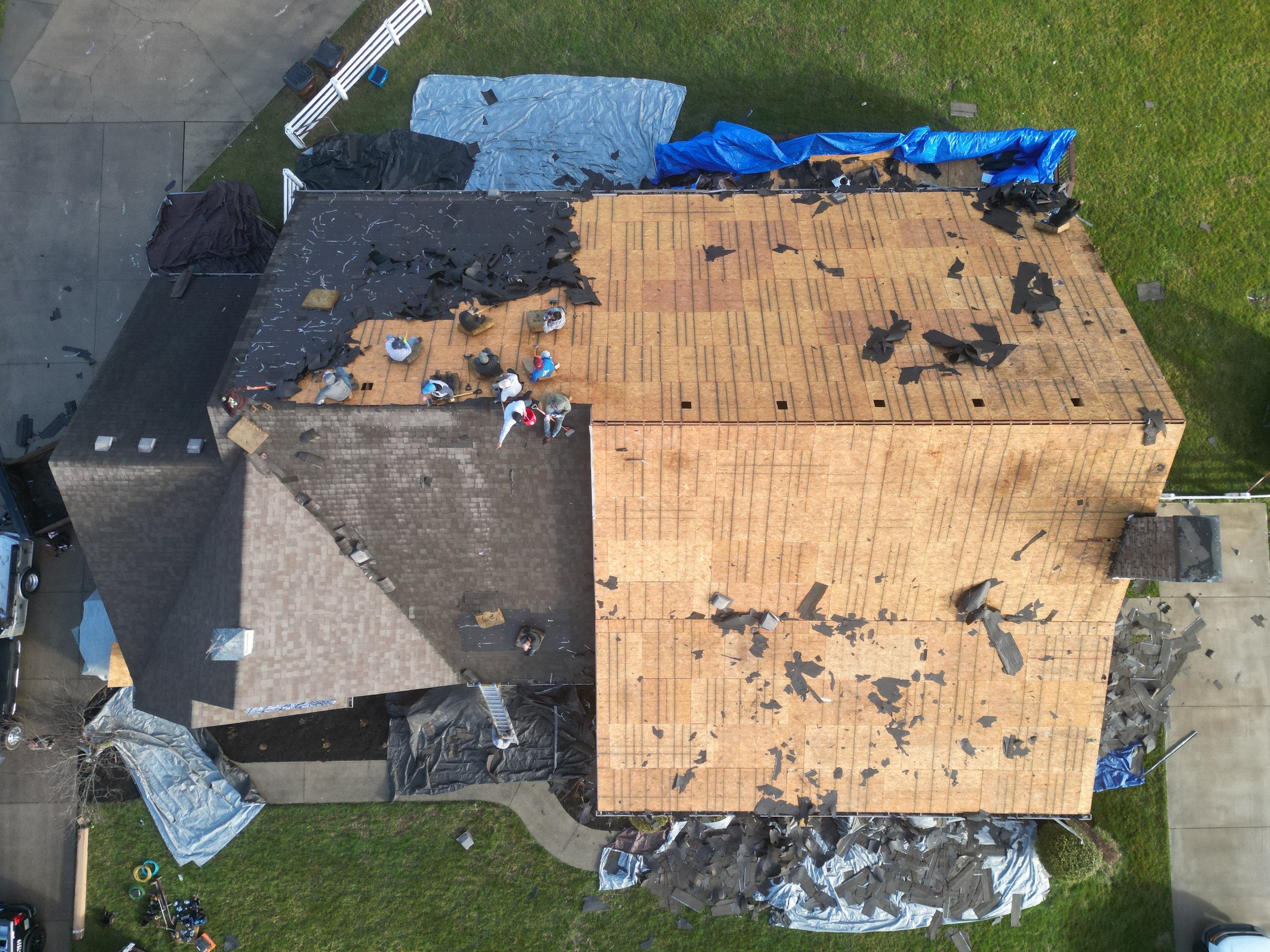 Aerial view of a house with its roof being replaced. The old asphalt shingle roof has been stripped away, exposing the wooden deck underneath. Workers are present on the roof, and tarps cover some areas of the house.