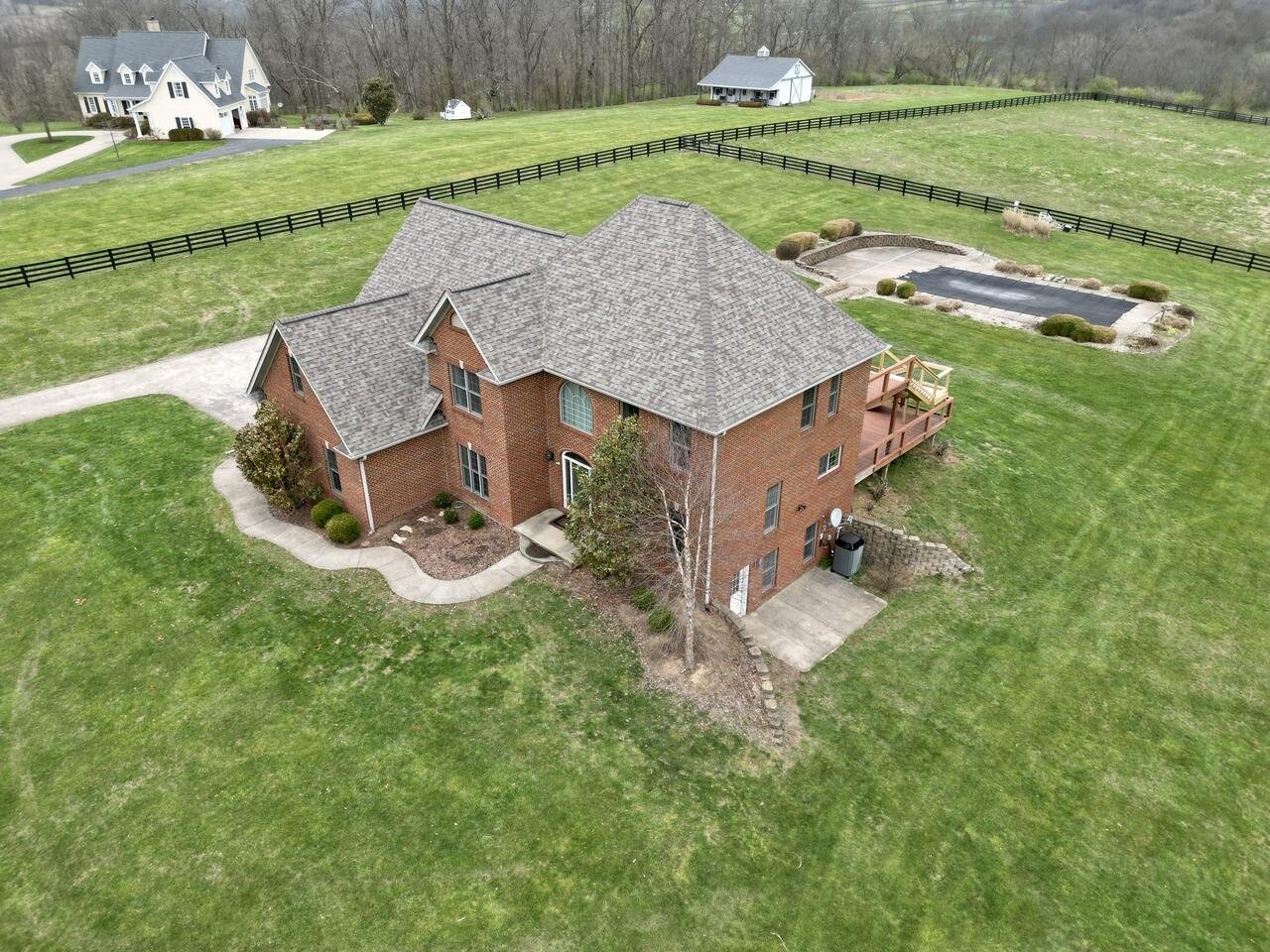 Aerial view of a large brick house with a landscaped backyard, deck, and fenced yard, surrounded by green grass and other houses in the distance.