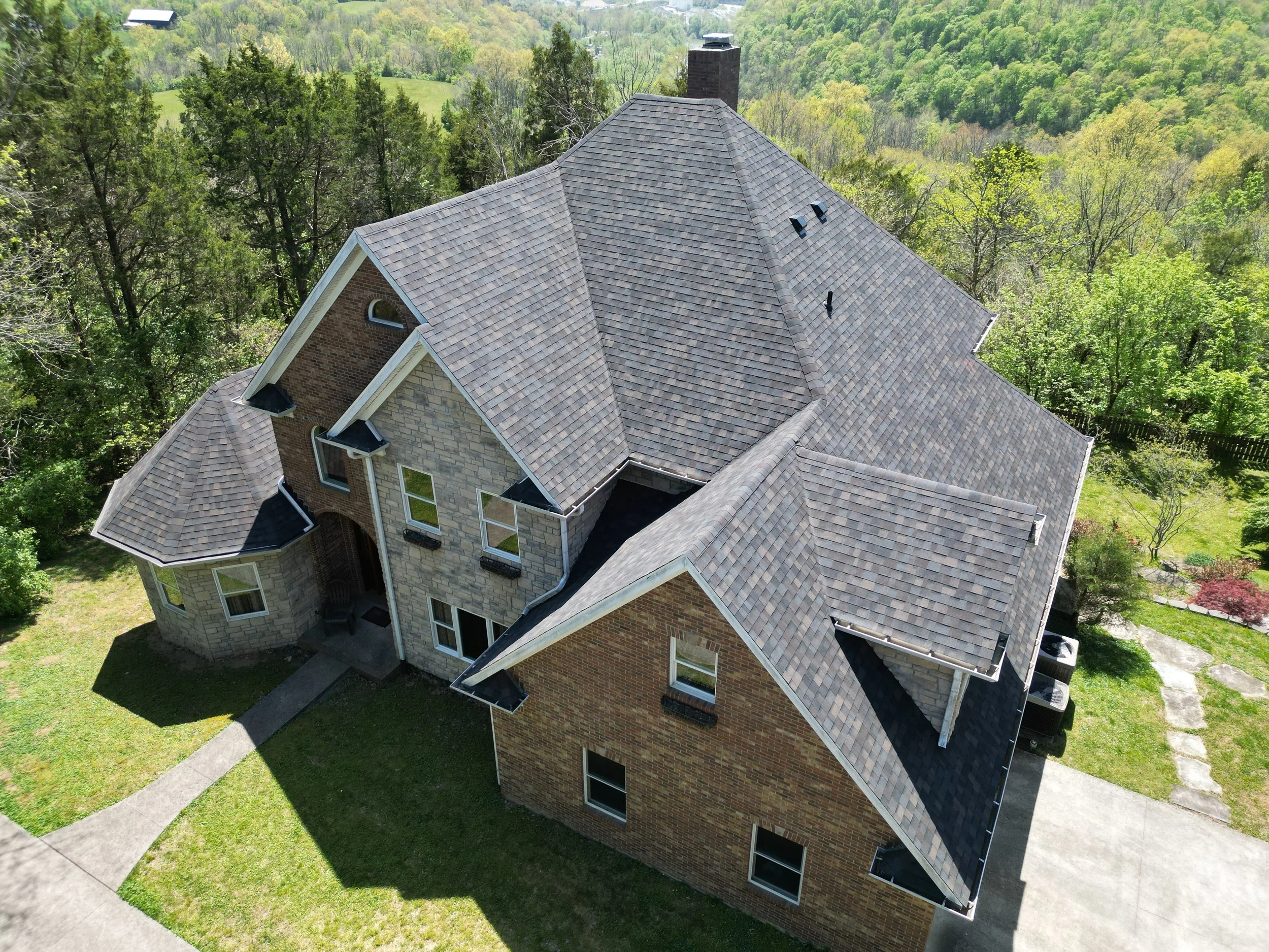 Aerial view of a large residential house with a multi-gabled roof, surrounded by green trees and a landscaped yard.
