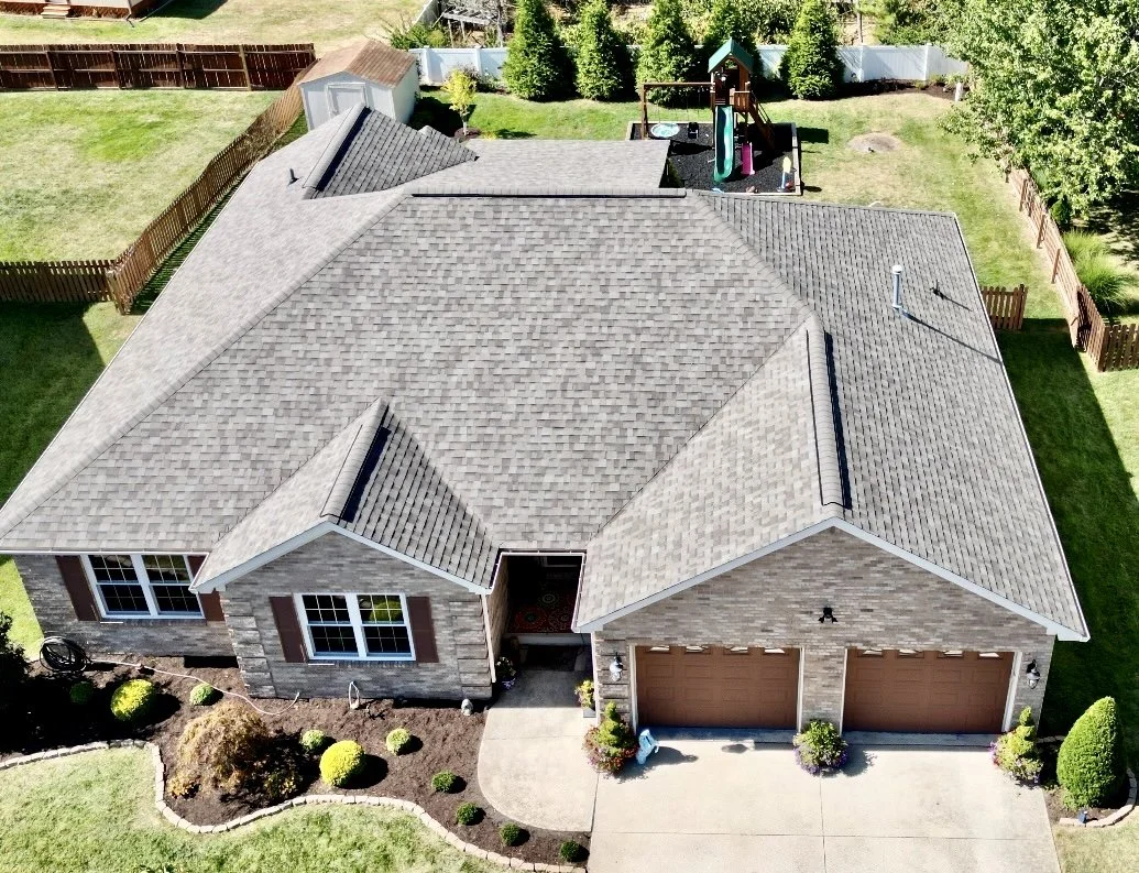 Aerial view of a single-story house with a gray shingle roof, brick exterior, and a two-car garage, surrounded by a lawn and fenced backyard with a children's playset.