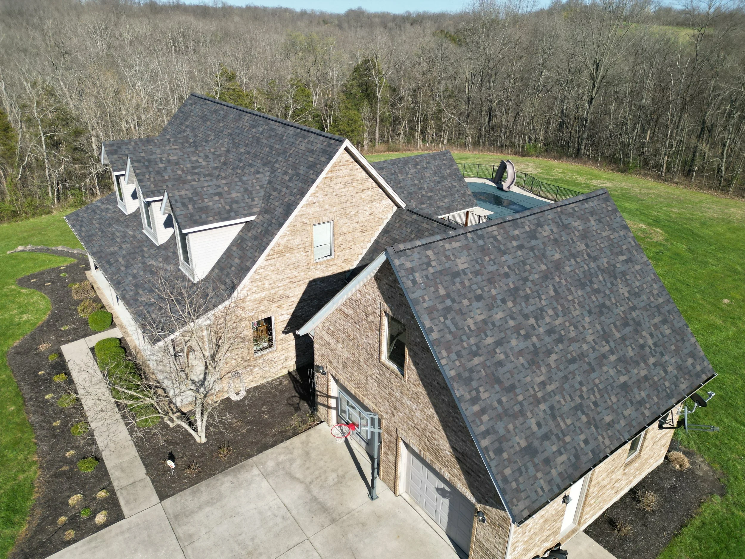 Aerial view of a suburban brick house with a multi-gabled roof, front porch, attached garage, landscaped yard, leafless trees, and a wooded area in the background.