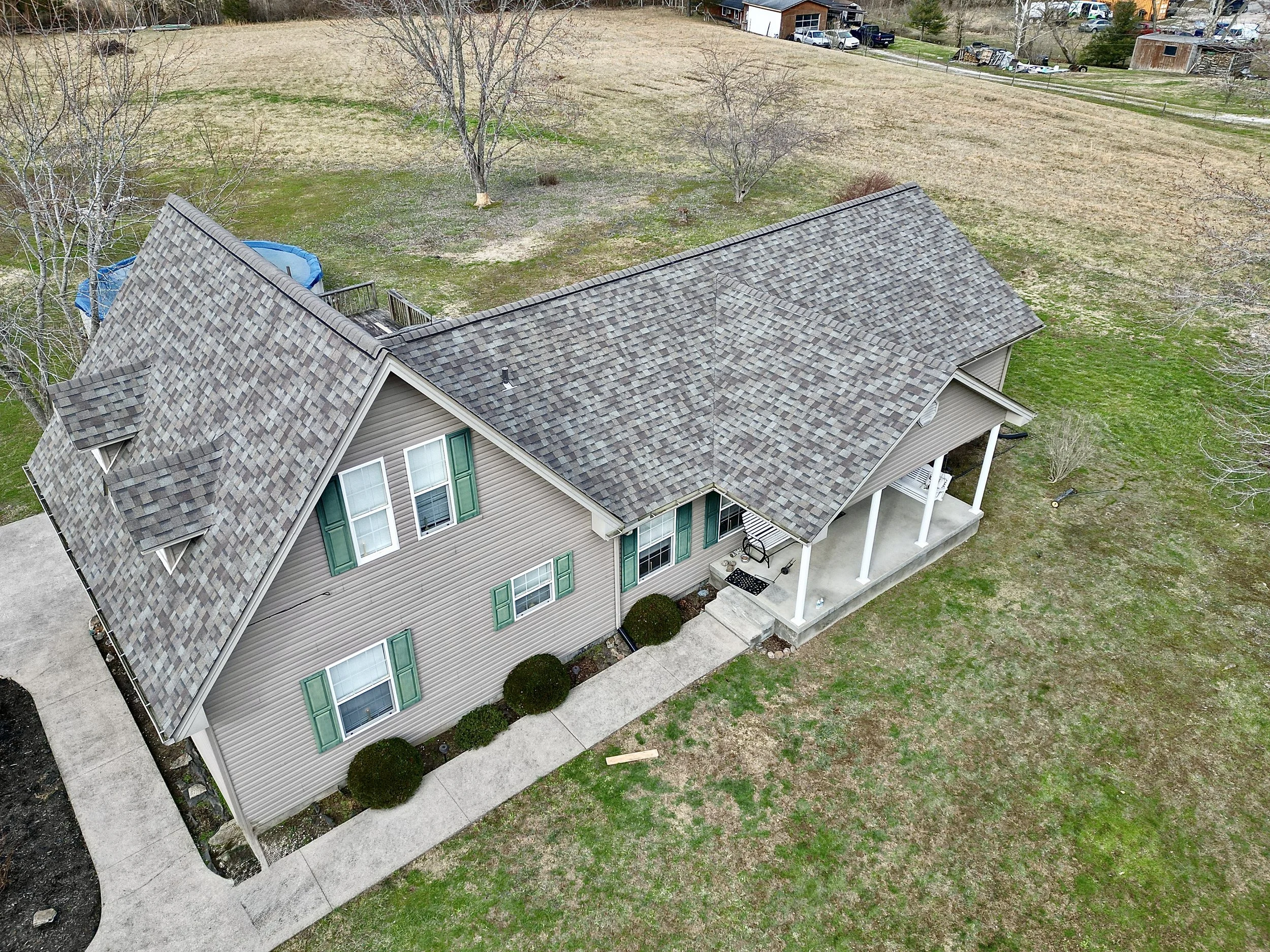 Aerial view of a house with beige siding, green shutters, a covered porch, a well-maintained yard with bushes, and a gray shingled roof. The yard is partially grassy with some patches of dirt, and there are trees and neighboring houses in the backgro