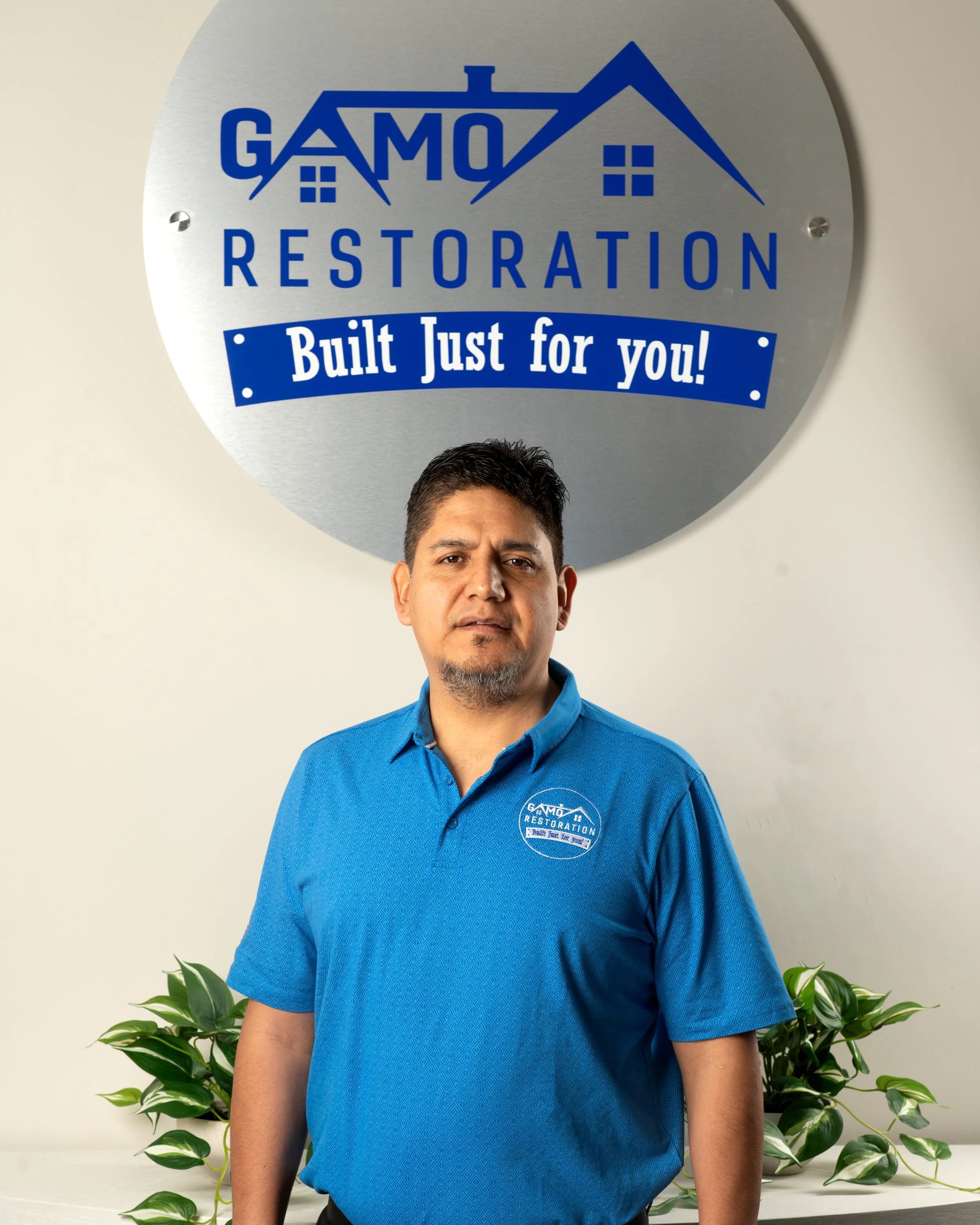 A man standing in front of a large circular sign that reads 'GAMO RESTORATION Built Just for you!' He is wearing a blue polo shirt with the company's logo on it, and there are green plants on the counter in front of him.