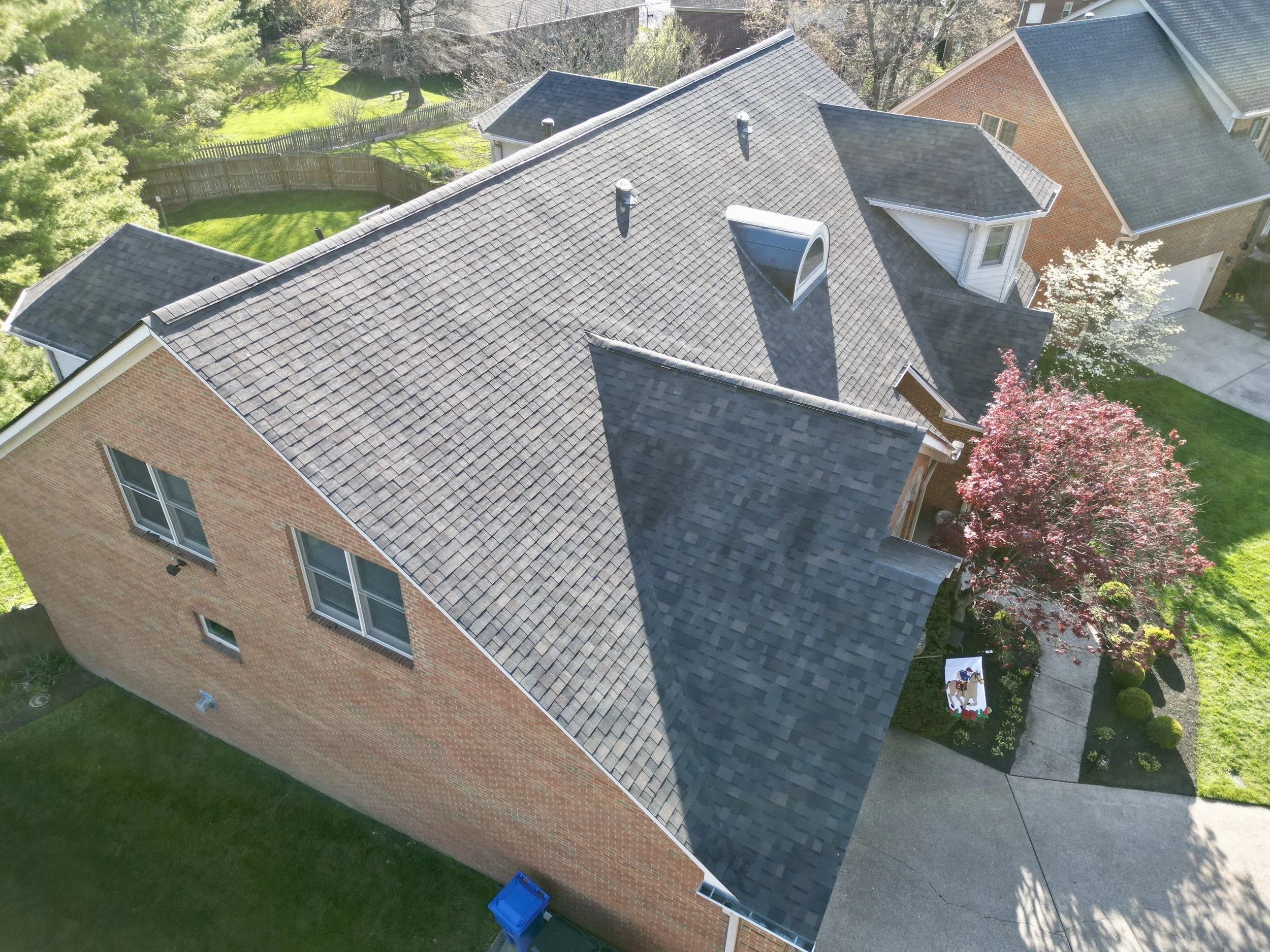 An aerial view of a brick house with multiple black shingled roofs, surrounded by green lawns, trees, and neighboring houses.