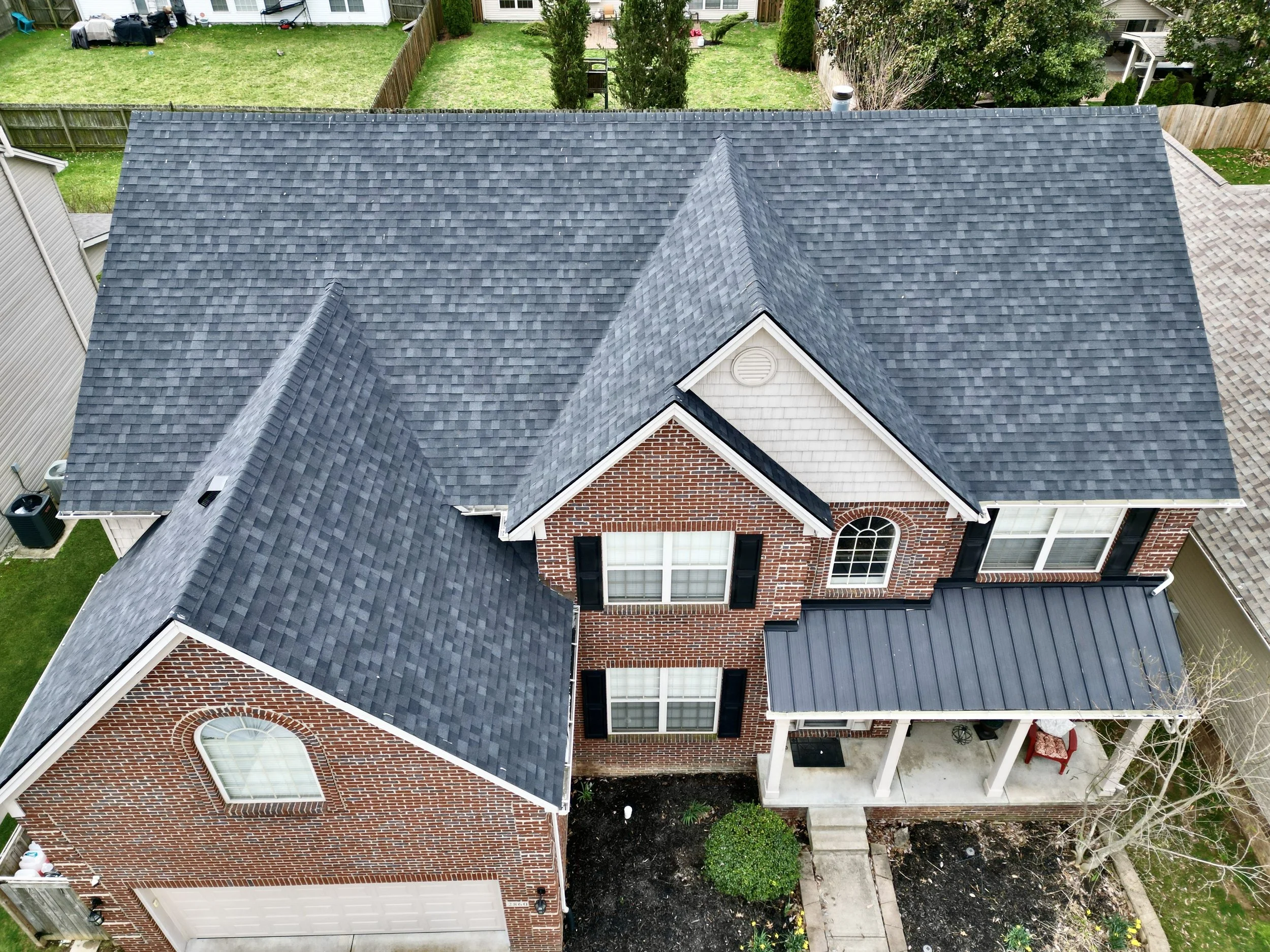 Aerial view of a brick house with a gray roof and a front porch with white columns, located in a backyard with a lawn and a garden.
