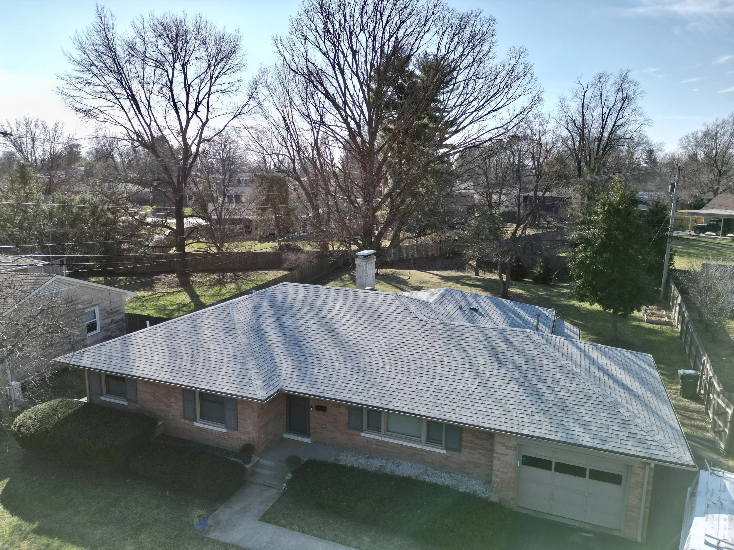 Aerial view of a suburban house with a grey shingle roof, brick exterior, and front yard with bushes and a concrete walkway, surrounded by trees and neighboring houses.