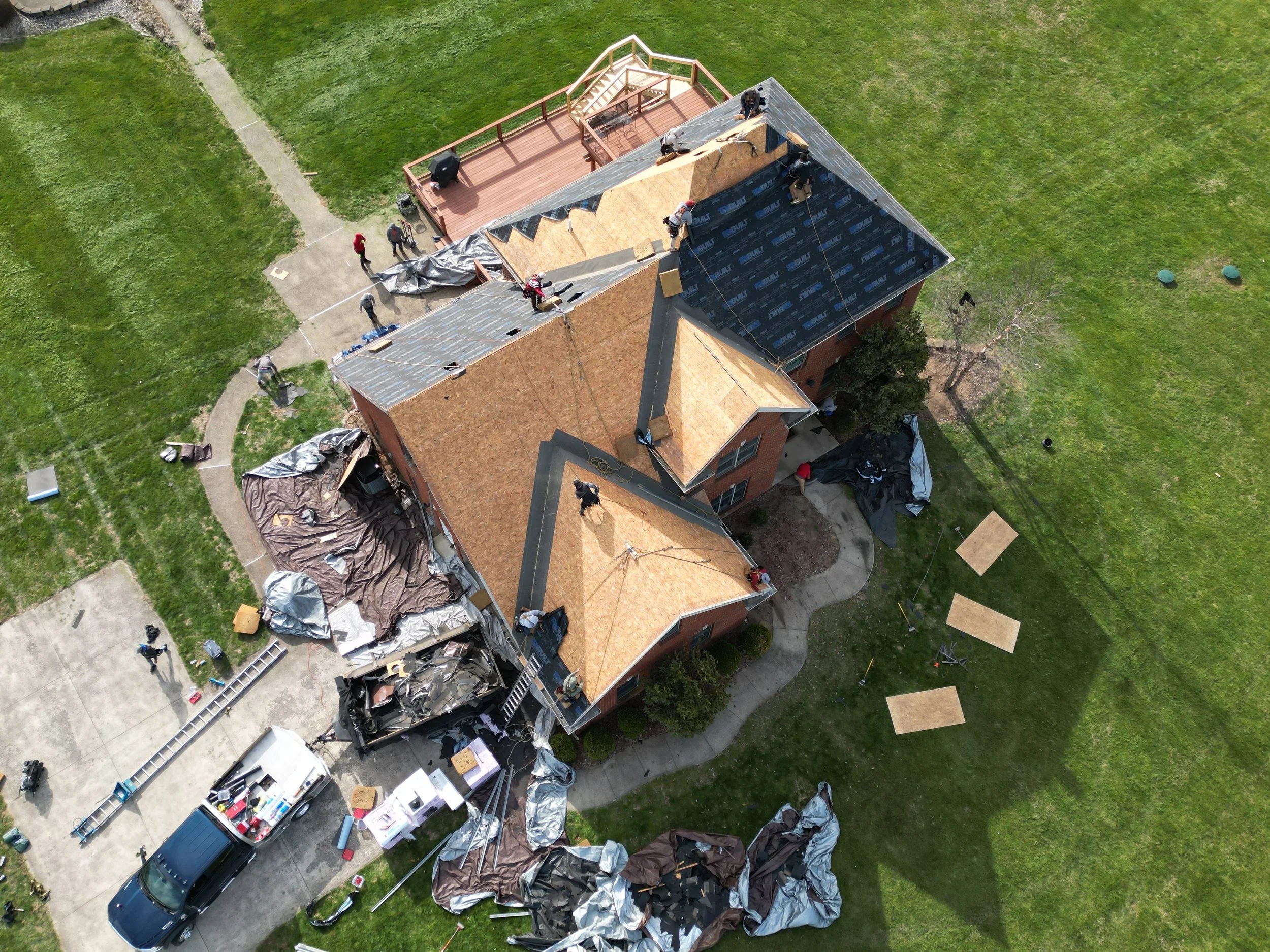 Aerial view of a house under construction with workers on the roof, covered in roofing materials, and equipment around the yard.