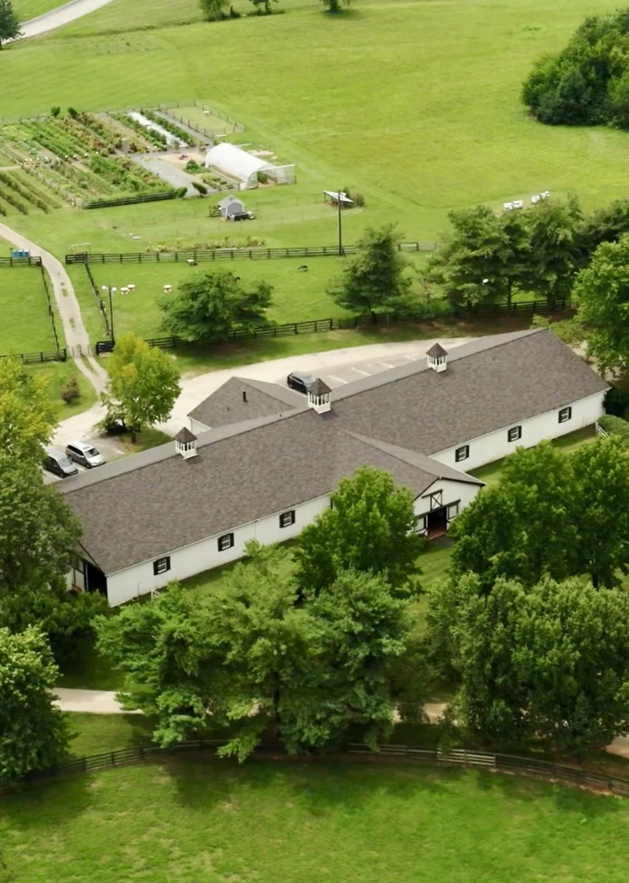 Aerial view of a farm with a long white barn, parking lot, green fields, trees, and a garden.