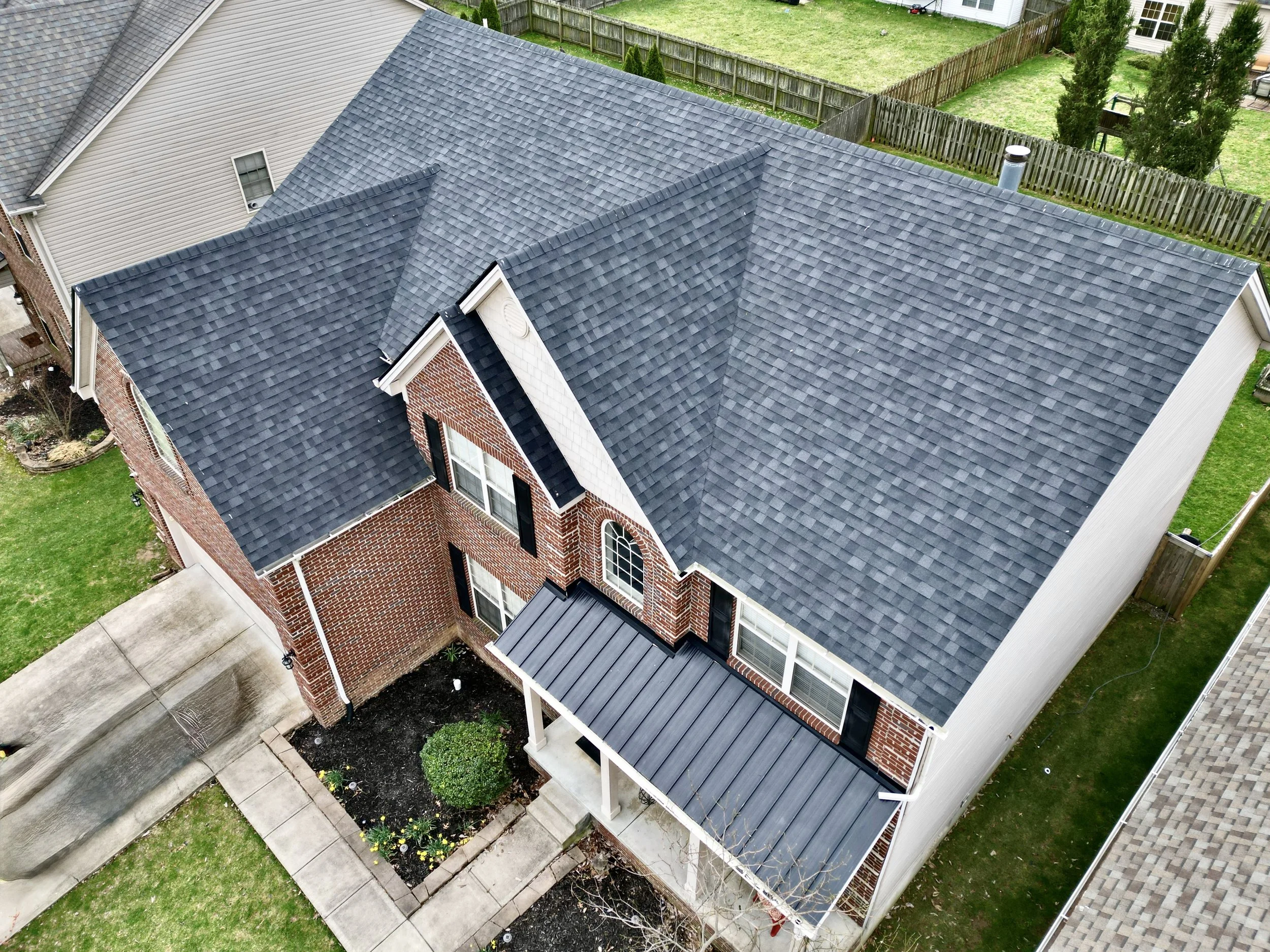 Aerial view of a house with a grey shingled roof, brick walls, and a small front porch with white columns and a metal roof, surrounded by a sidewalk, garden bed, and lawn.