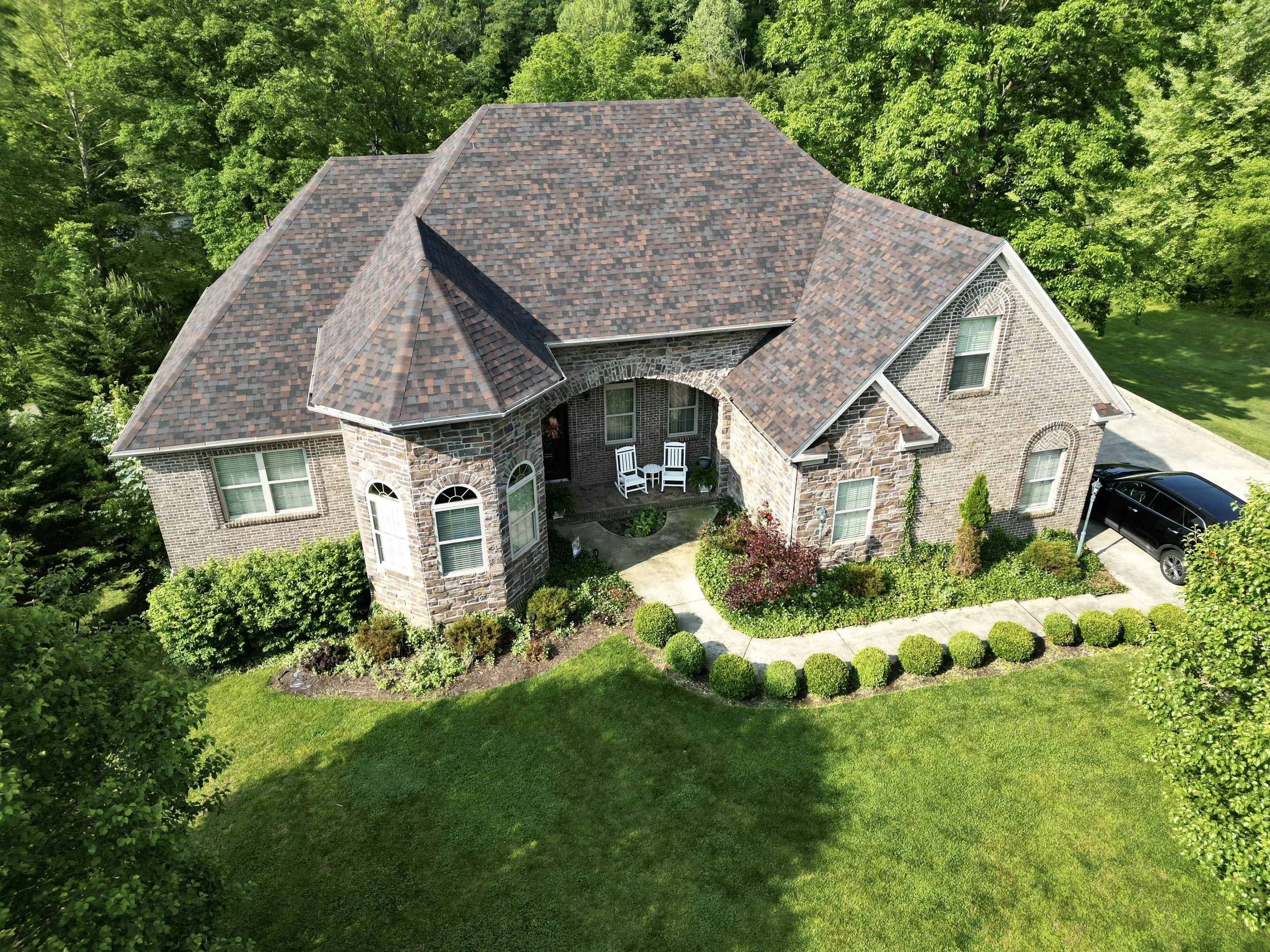 A large brick house with a complex roof, front porch with white chairs, surrounded by well-maintained lawn and bushes, black car parked on the driveway, and trees in the background.