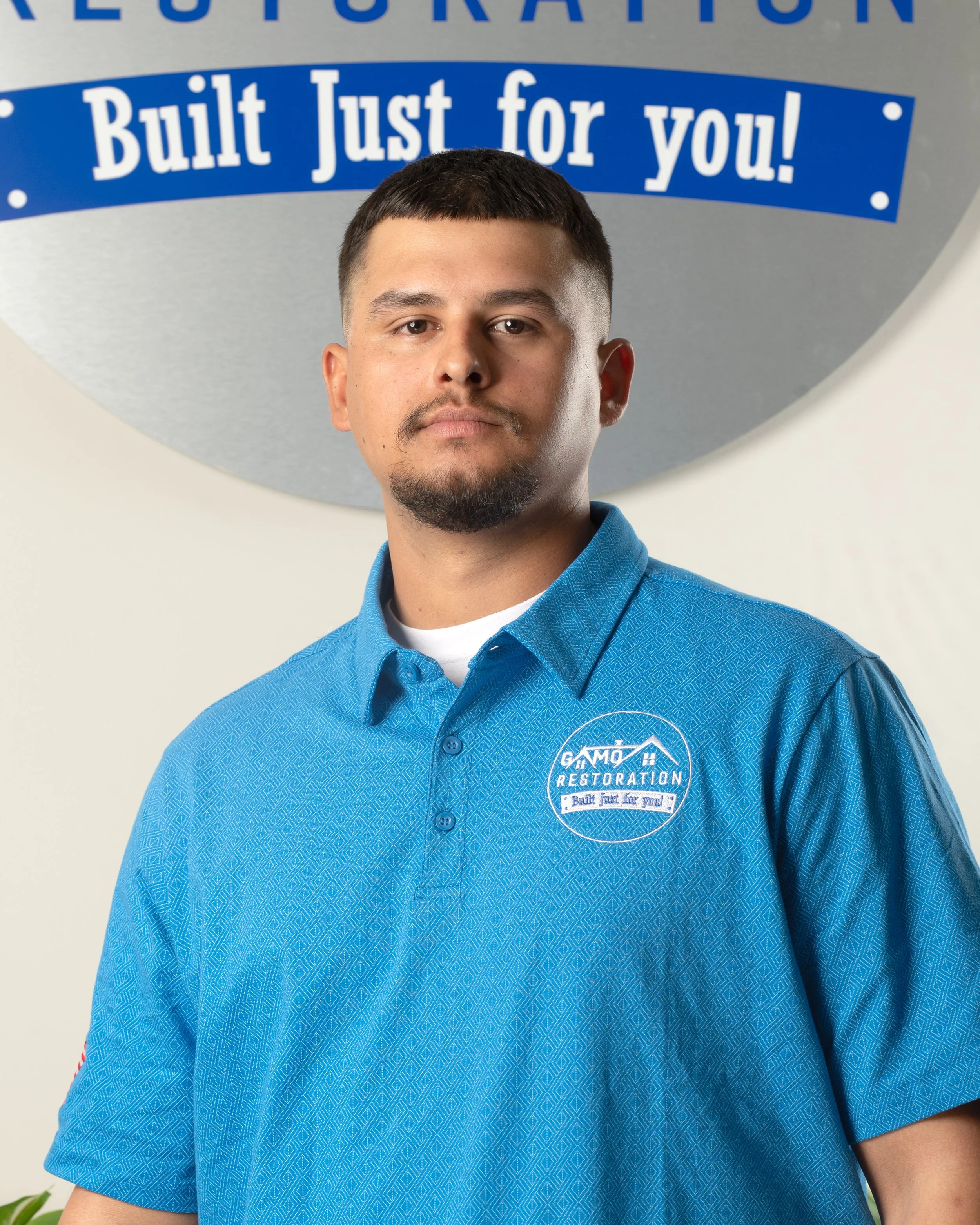 A man in a blue collared shirt standing in front of a circular sign that reads 'Built Just for You!' and 'Restoration'.