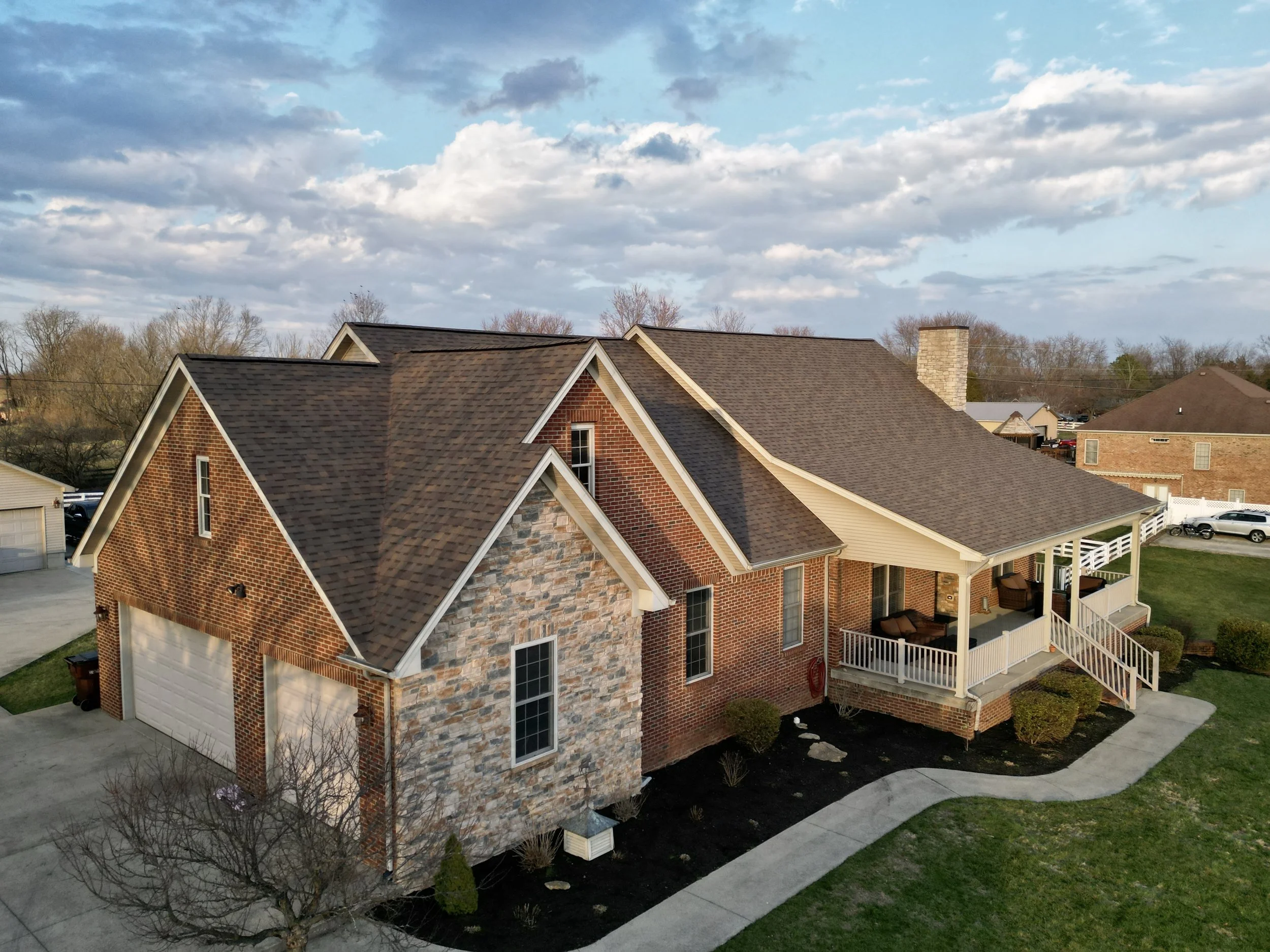 Aerial view of a brick house with a stone facade and a covered porch, surrounded by a lawn with a small tree and bushes, under a partly cloudy sky.