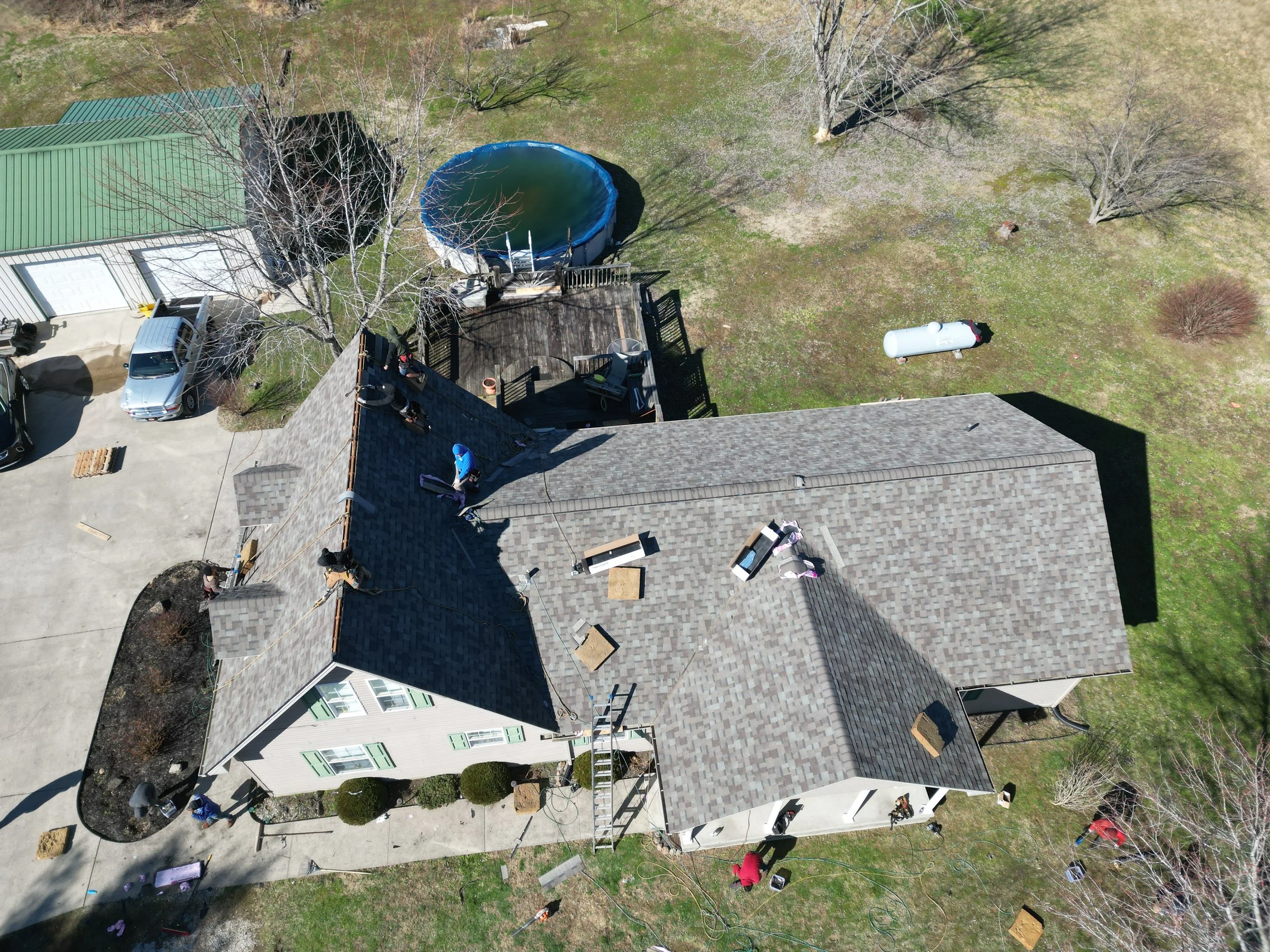 Aerial view of a house with roofing work being done by multiple workers. The house has a gray shingle roof, a ladder, and a deck at the back. There is a concrete driveway with a silver vehicle, a trampoline, a propane tank, and nearby trees with no l