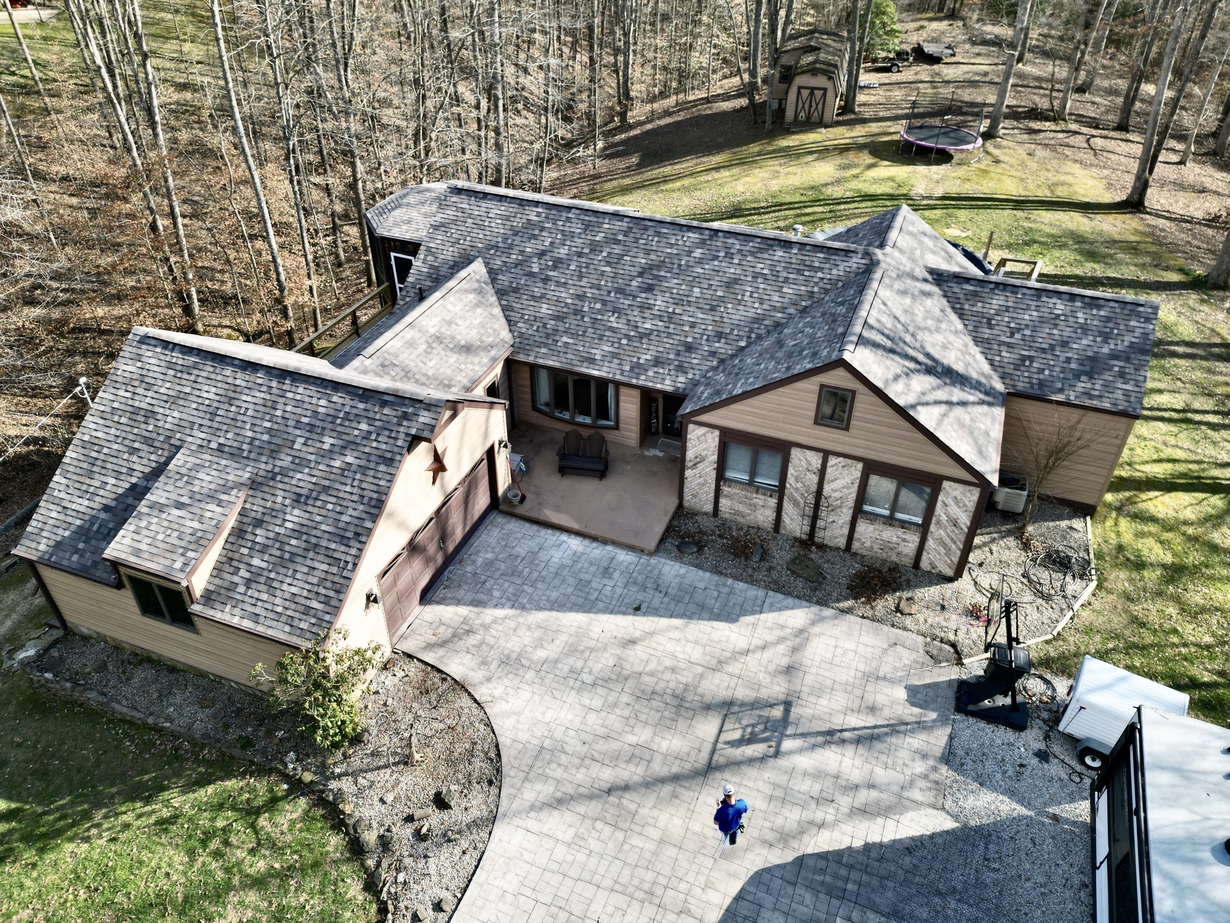 Aerial view of a house with a complex roof, attached garage, driveway, a small patio with seating, and a yard with a trampoline and trees.