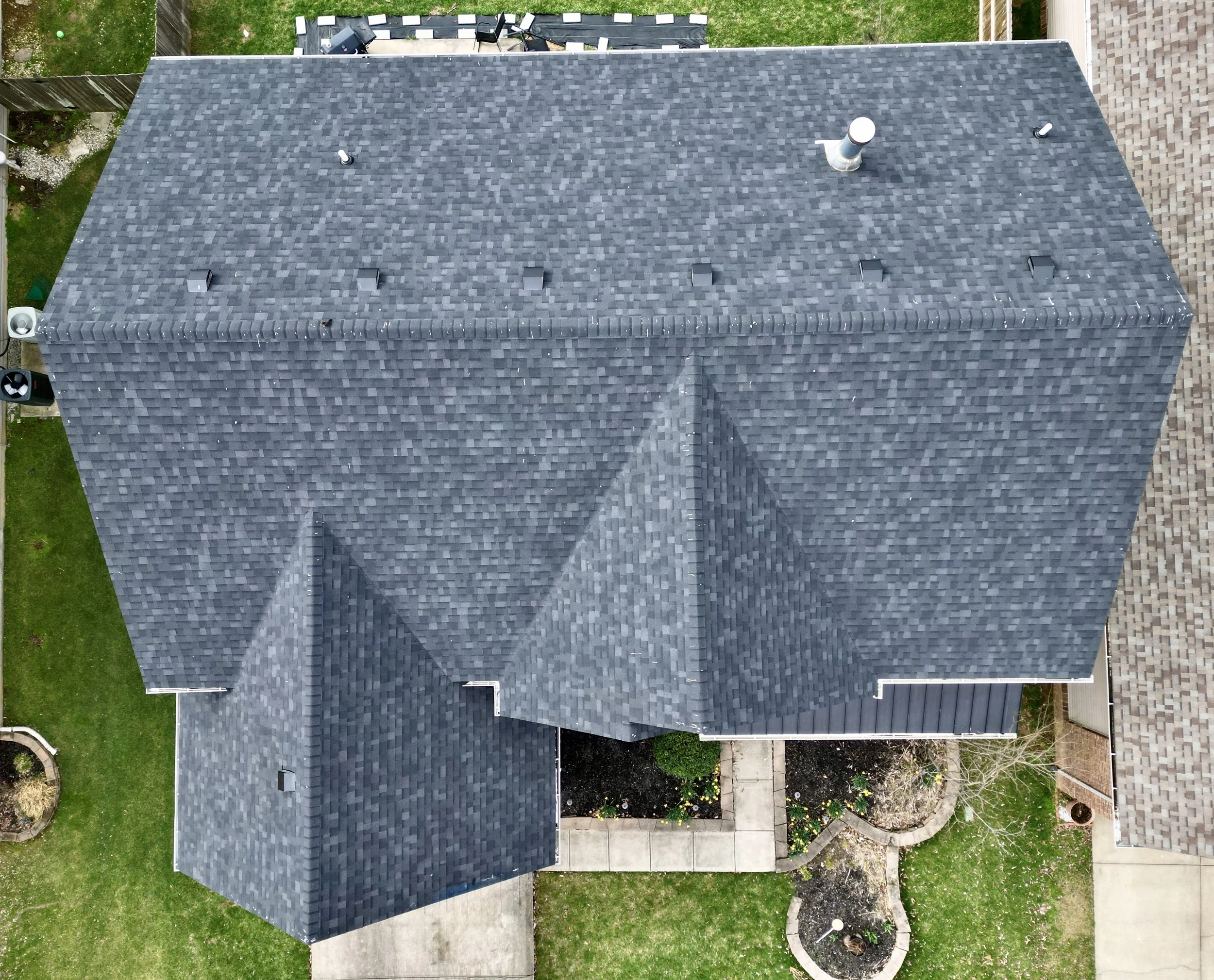 Aerial view of a house with a gray shingled roof, surrounded by a green lawn, concrete walkways, and landscaped garden beds.