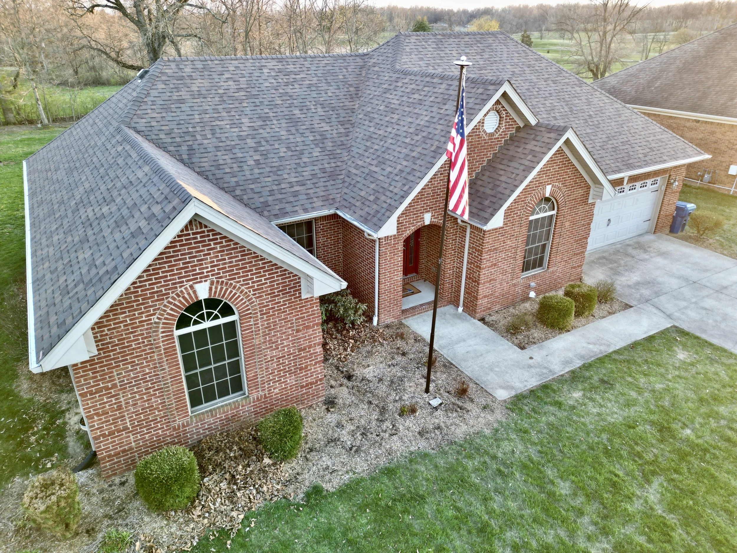 An aerial view of a one-story brick house with a gable roof, front porch, American flag, and a concrete driveway leading to an attached two-car garage, surrounded by green lawn and trees.