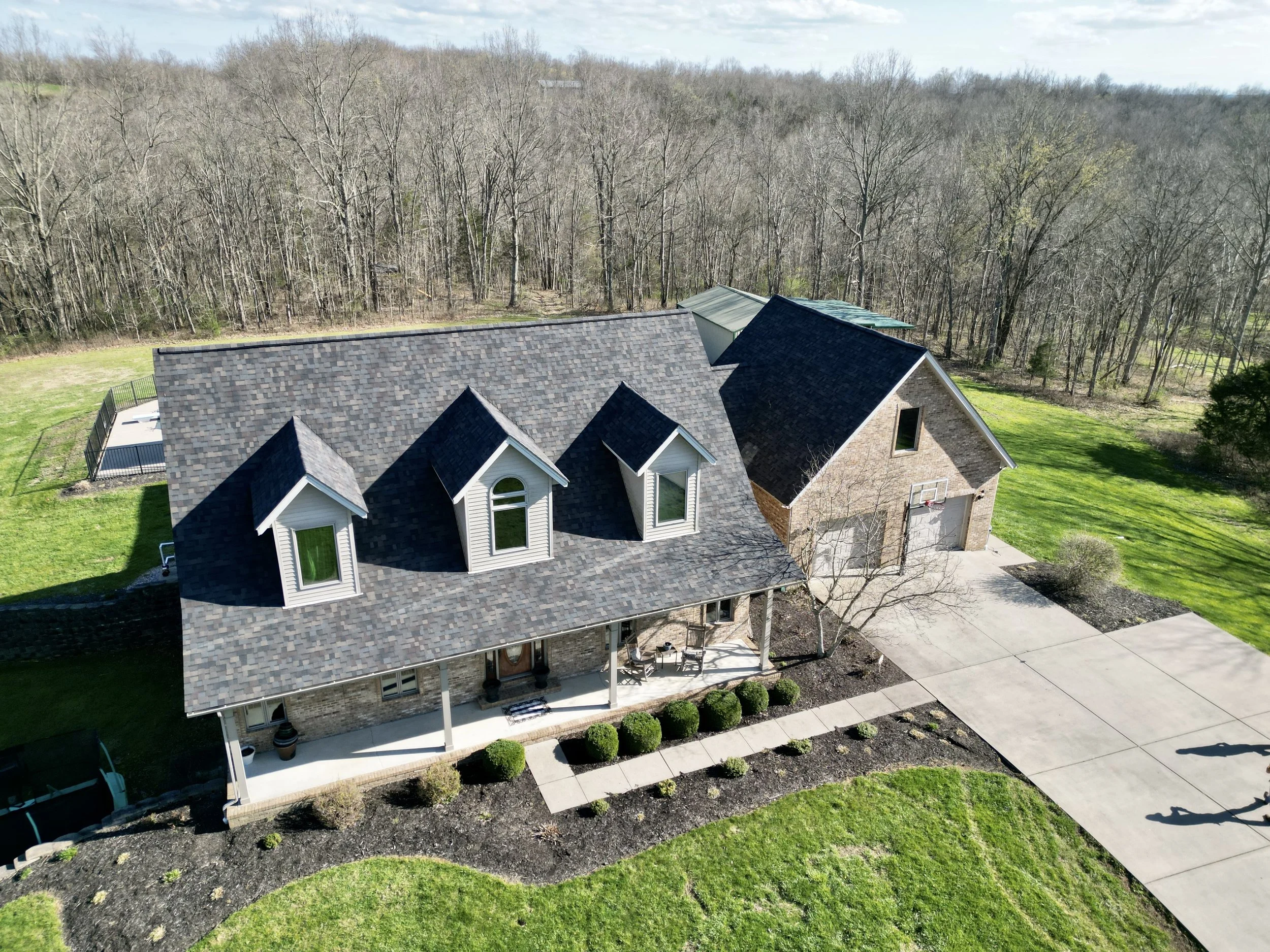 Aerial view of a large house with a brick and siding exterior, a multi-gabled roof with three dormer windows, a front porch with chairs, a driveway, a fenced backyard with a patio, and surrounding green lawn and trees.