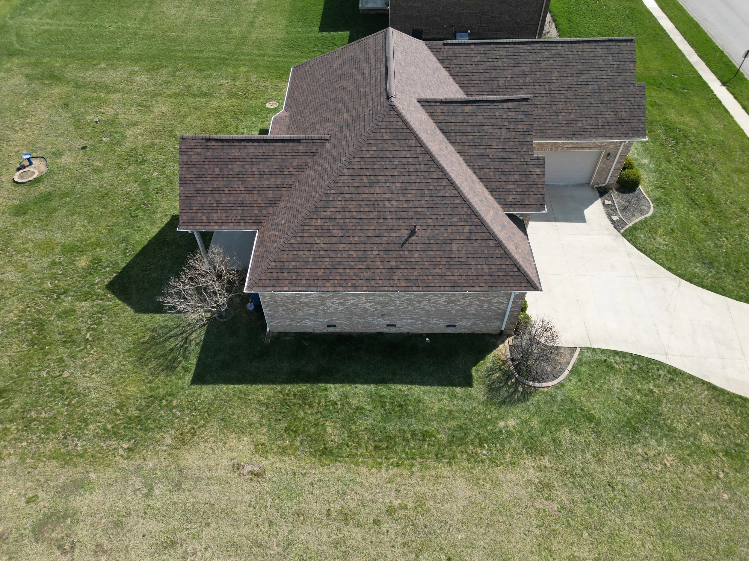 Aerial view of a house with a brown shingled roof, brick exterior walls, a driveway, and a lawn with trees.