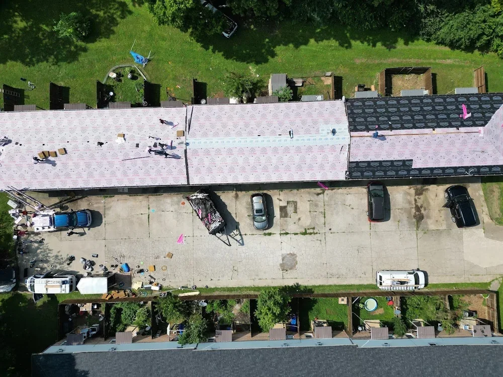 An aerial view of a building under construction with workers installing roofing materials. A parking lot with several parked cars and construction equipment is visible below. There are trees and backyard structures surrounding the scene.