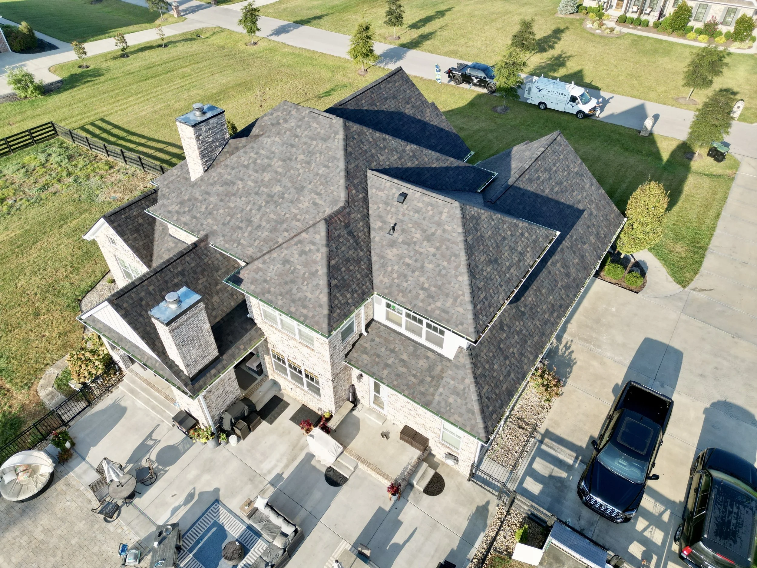 Aerial view of a large house with multiple gabled roofs, brick and siding exterior, and a spacious backyard with patio furniture, surrounded by a green lawn and trees. Several vehicles are parked on the driveway and street.