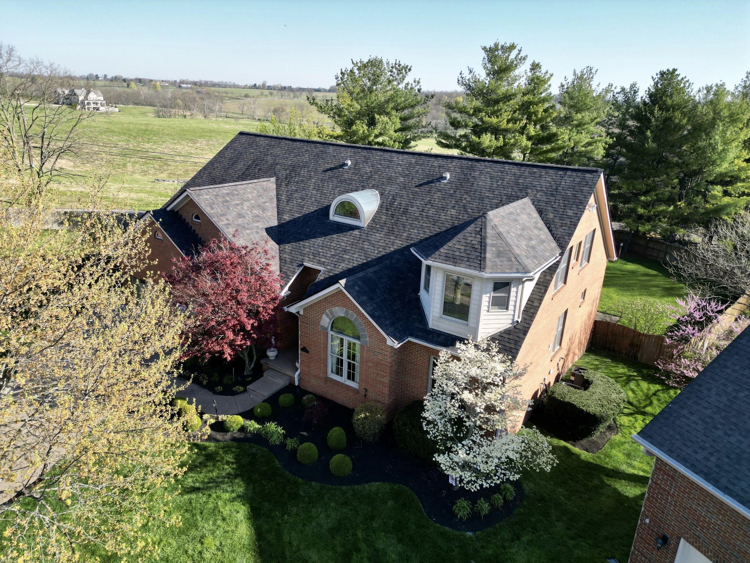 Aerial view of a brick house with a dark shingle roof and white trim, surrounded by trees with blooming flowers, in a green yard with a curved pathway and private backyard fencing, with open fields and distant houses under a clear sky.