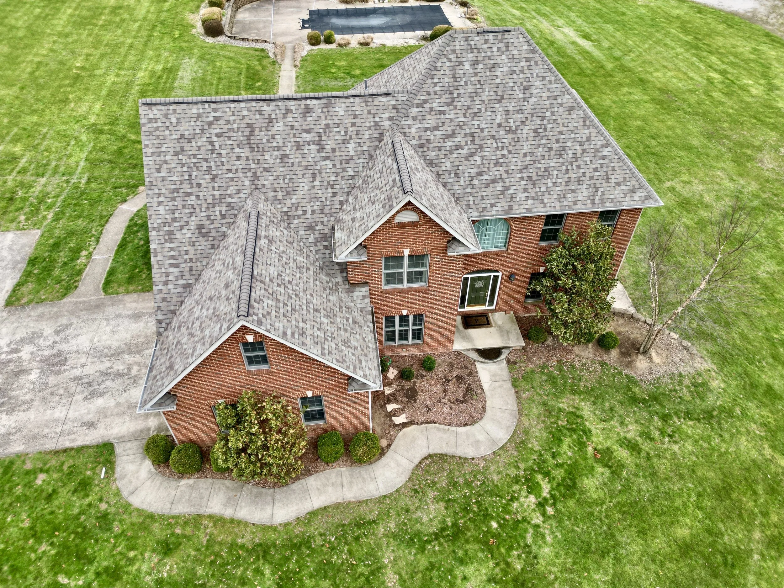 Aerial view of a brick house with a gray shingled roof, surrounded by green grass, trees, and a curved concrete sidewalk leading to the front entrance.