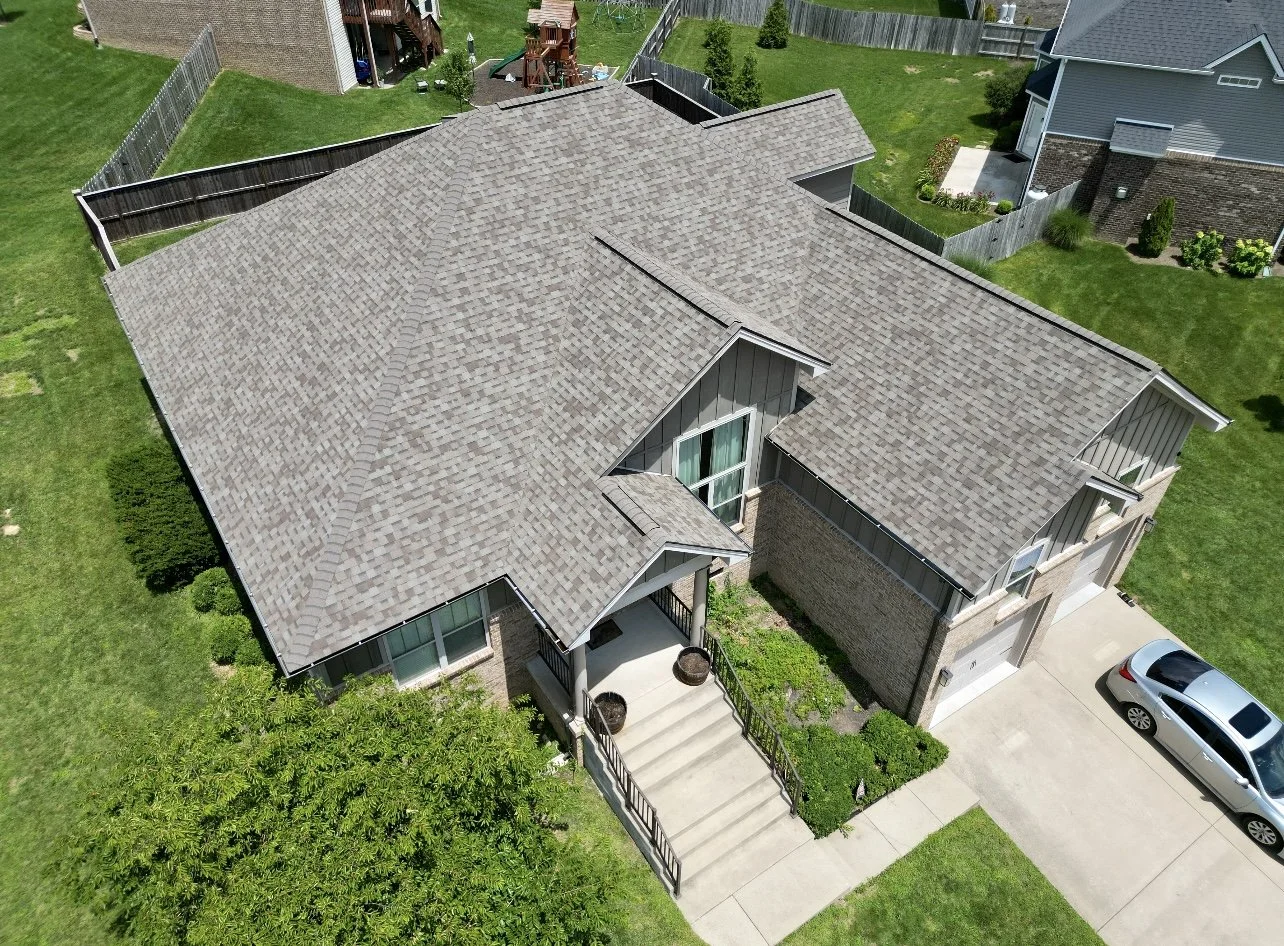 An aerial view of a suburban house with a large, multi-gabled shingled roof, front porch with steps, and a driveway with a silver car. The house is surrounded by a well-maintained lawn and other houses with similar yards.