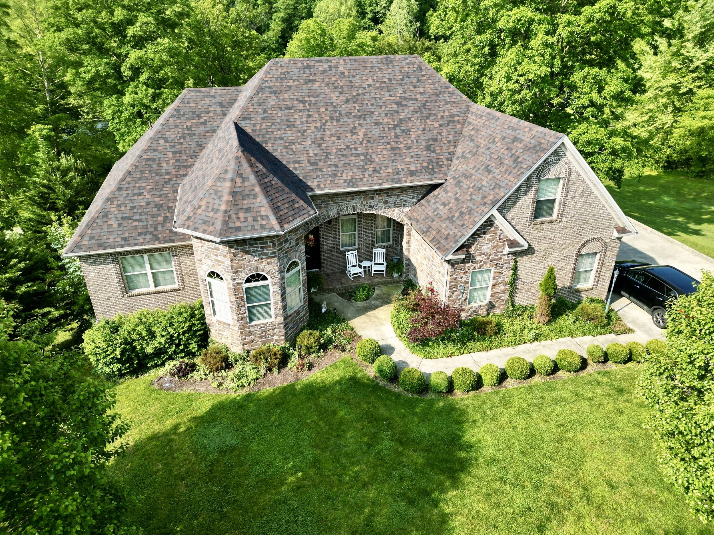 Aerial view of a large brick house with a brown shingle roof, surrounded by green trees and a well-maintained lawn with garden beds and shrubs.