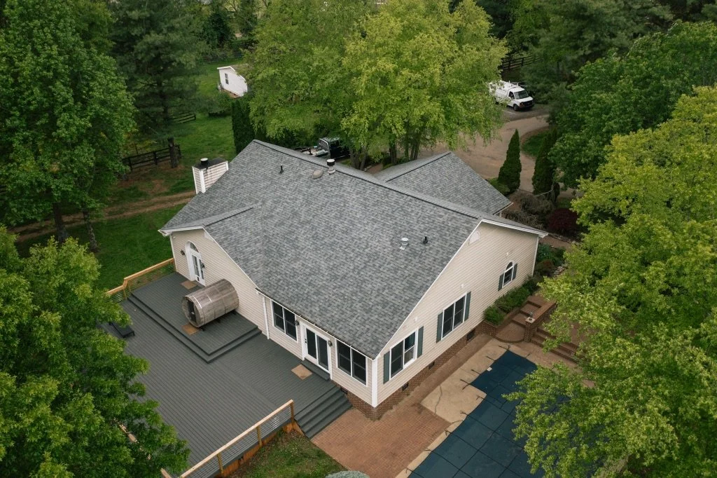 Aerial view of a beige house with a gray roof, surrounded by green trees and a backyard with a deck, patio, and outdoor furniture.