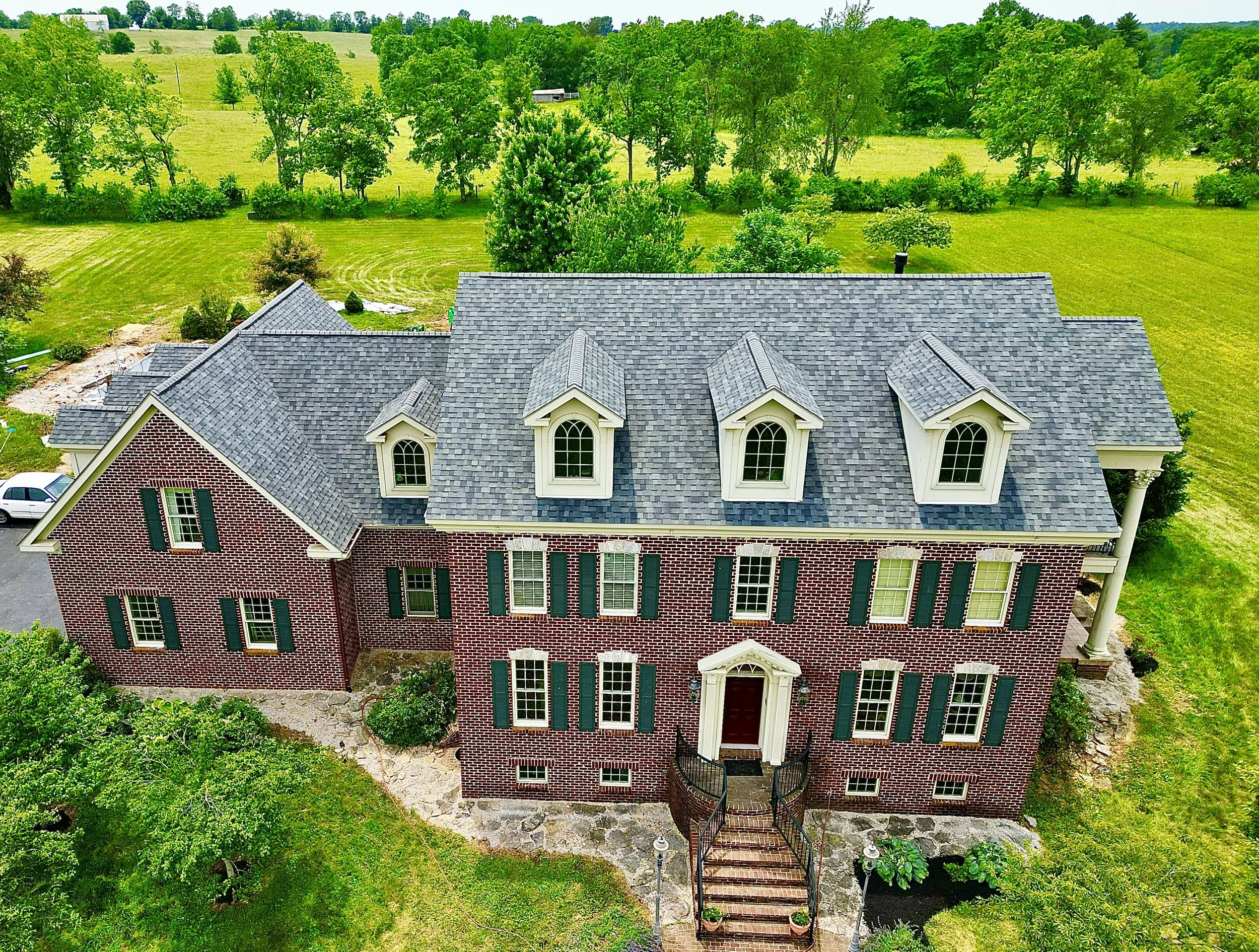 A large brick house with a gray shingle roof, multiple dormer windows, and green shutters, surrounded by a lush green lawn and trees.