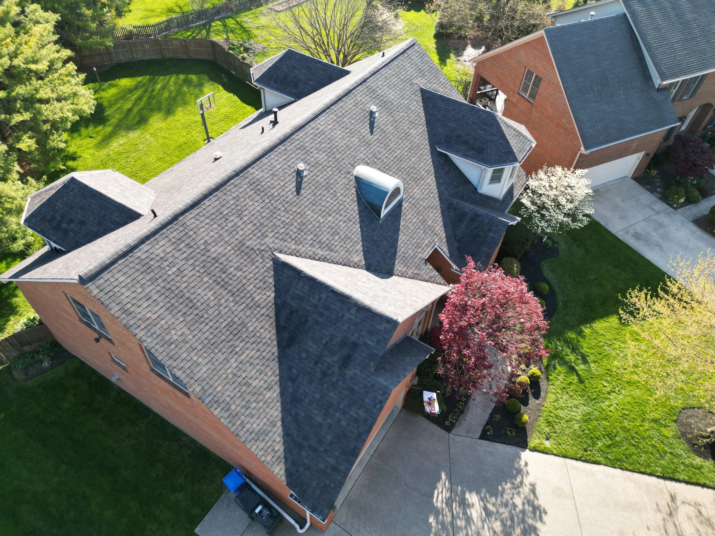 An aerial view of a suburban house with a dark shingled roof, brick exterior, and well-maintained yard with green grass, trees, and a concrete driveway.