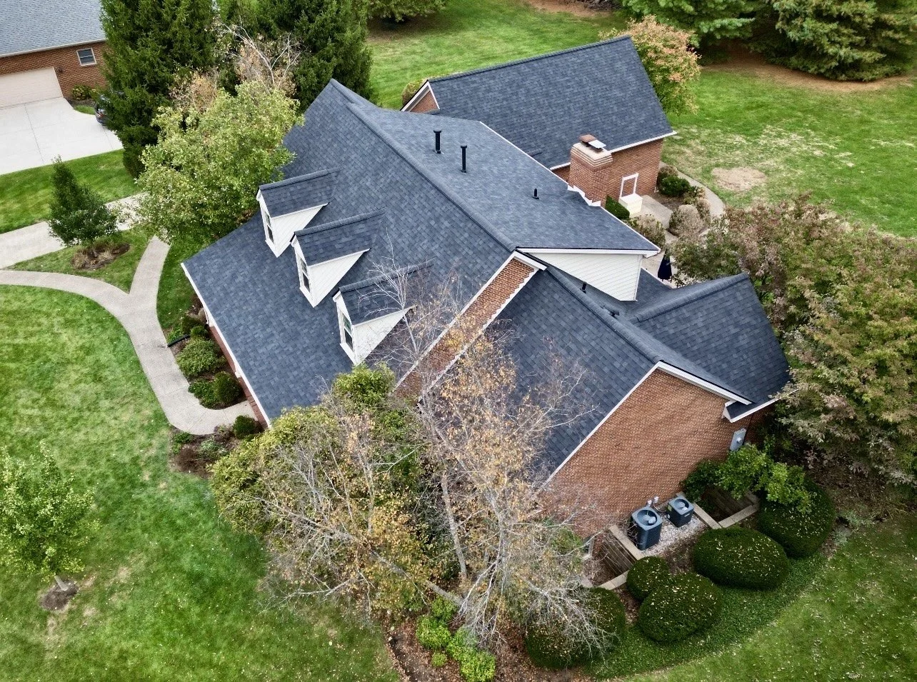 An aerial view of a house with a dark shingled roof, surrounded by green grass and trees, with a paved walkway leading to the front door.