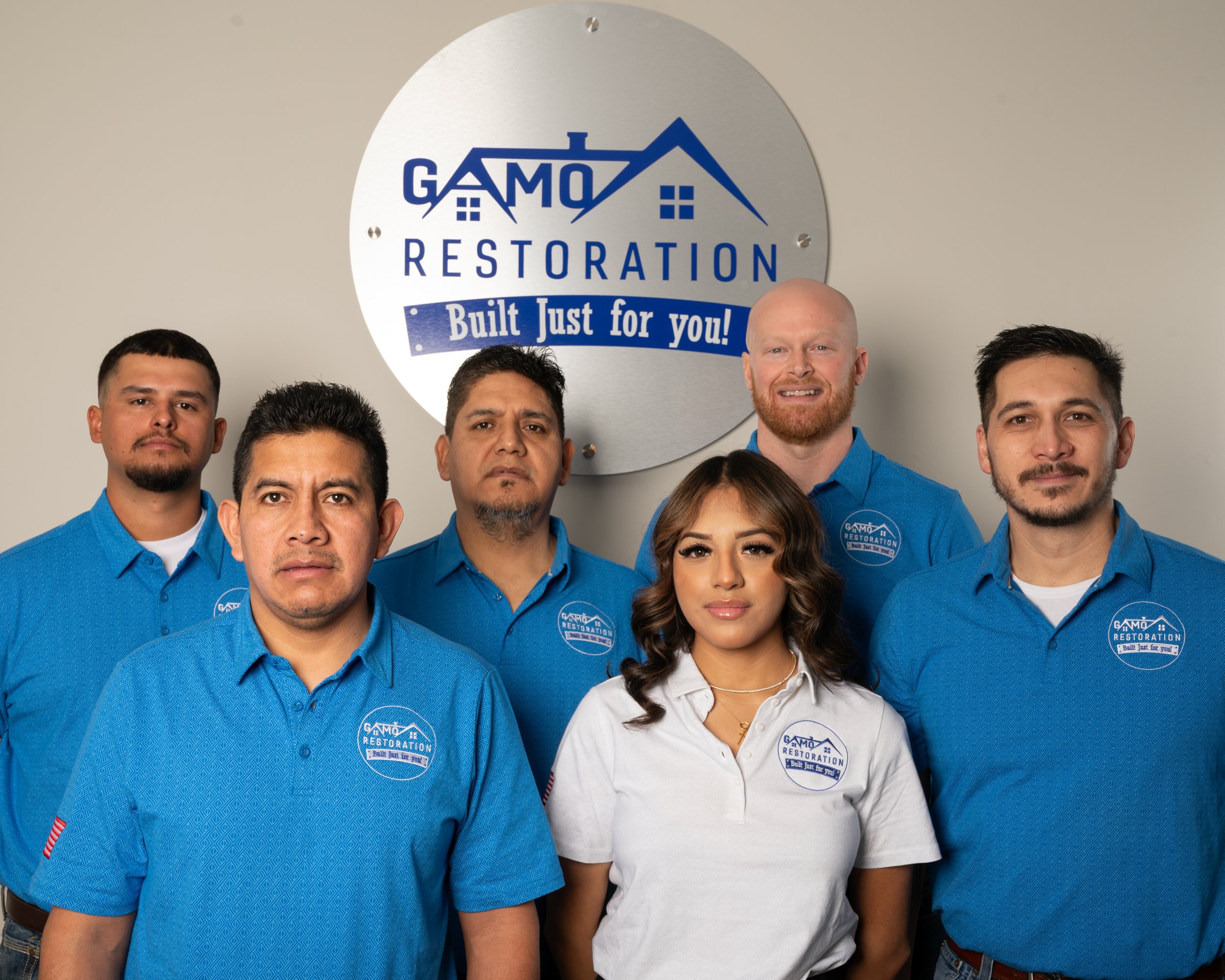 A group of six employees wearing blue and white uniforms with the Gamo Restoration logo, standing in front of a Gamo Restoration sign on a gray wall.