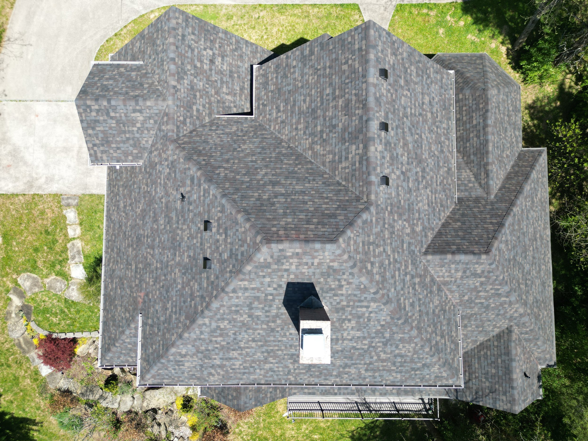 An aerial view of a house with a complex, multi-gabled gray shingle roof, surrounded by a yard with grass, trees, and a concrete driveway.