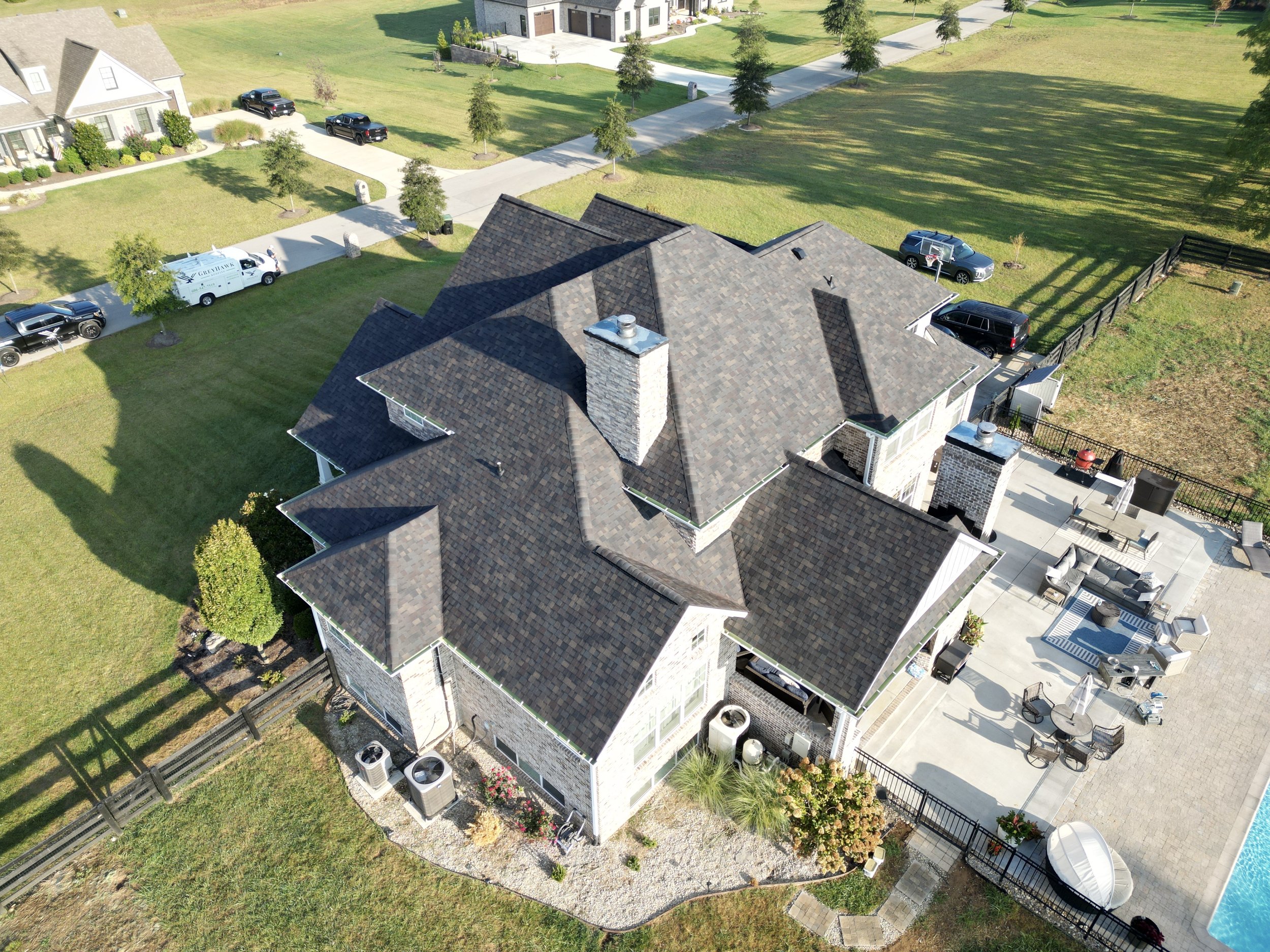 Aerial view of a large suburban house with a dark shingle roof, a spacious backyard with a patio and outdoor furniture, surrounded by a black fence and neighboring houses. Green lawns and trees are also visible.