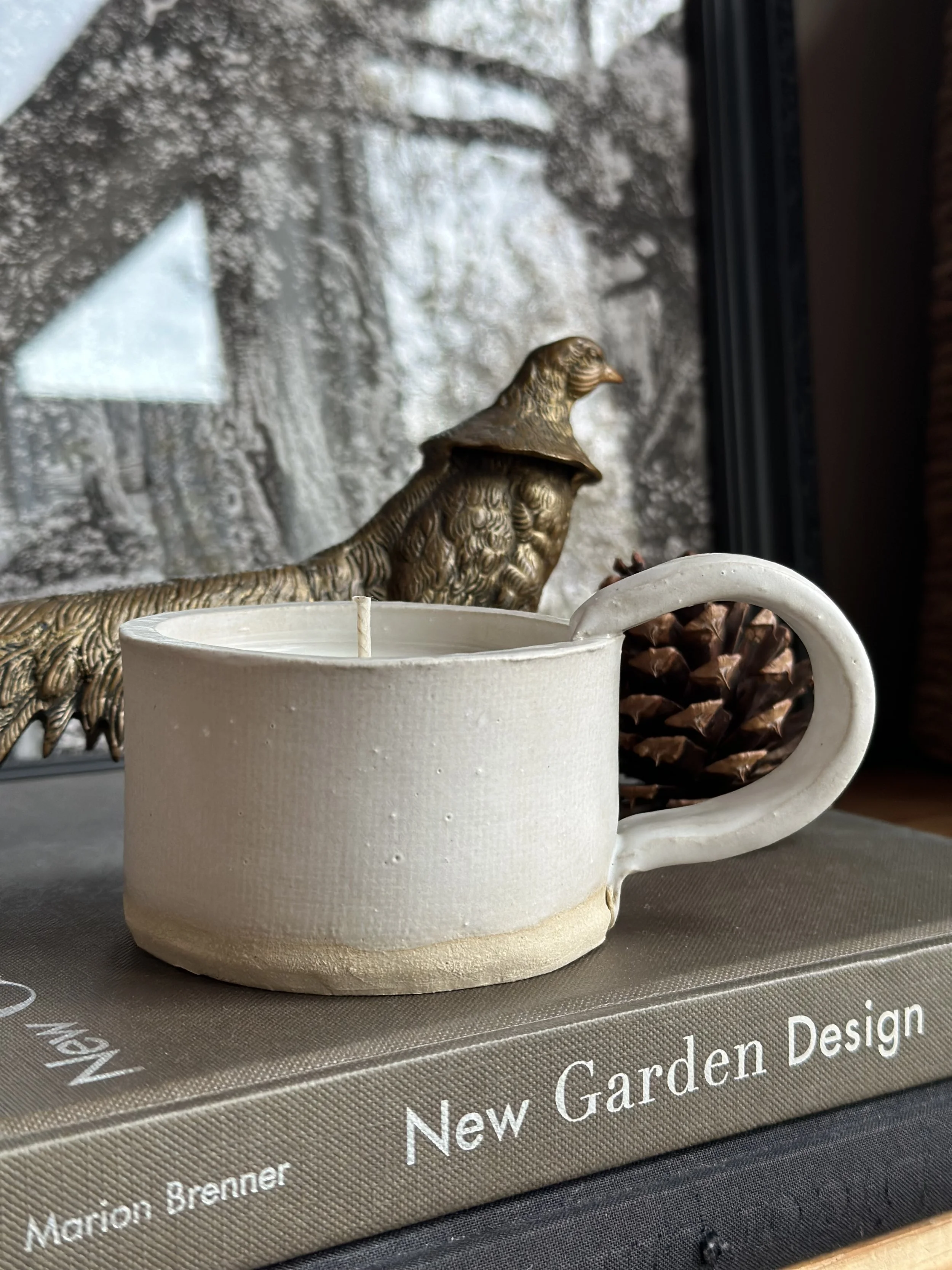 Decorative ceramic mug with a pinecone, a gold-colored bird figurine, a book titled 'New Garden Design' by Marion Brenner, and a window with a snow-covered landscape outside.