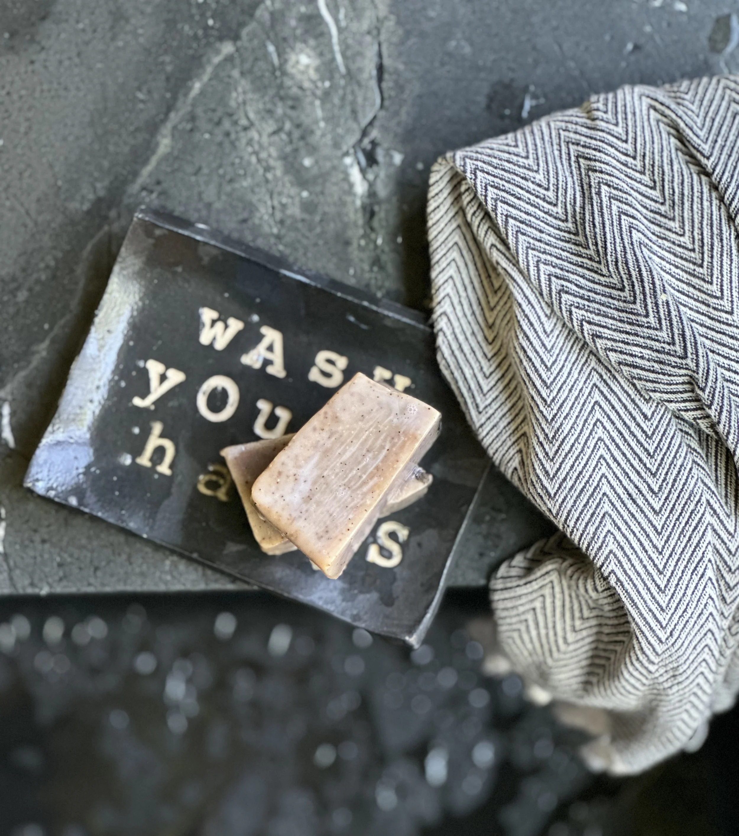 A soap bar placed on a black, distressed surface with the words 'WASH' and 'YOU' partially visible. A striped cloth is draped to the right of the soap.
