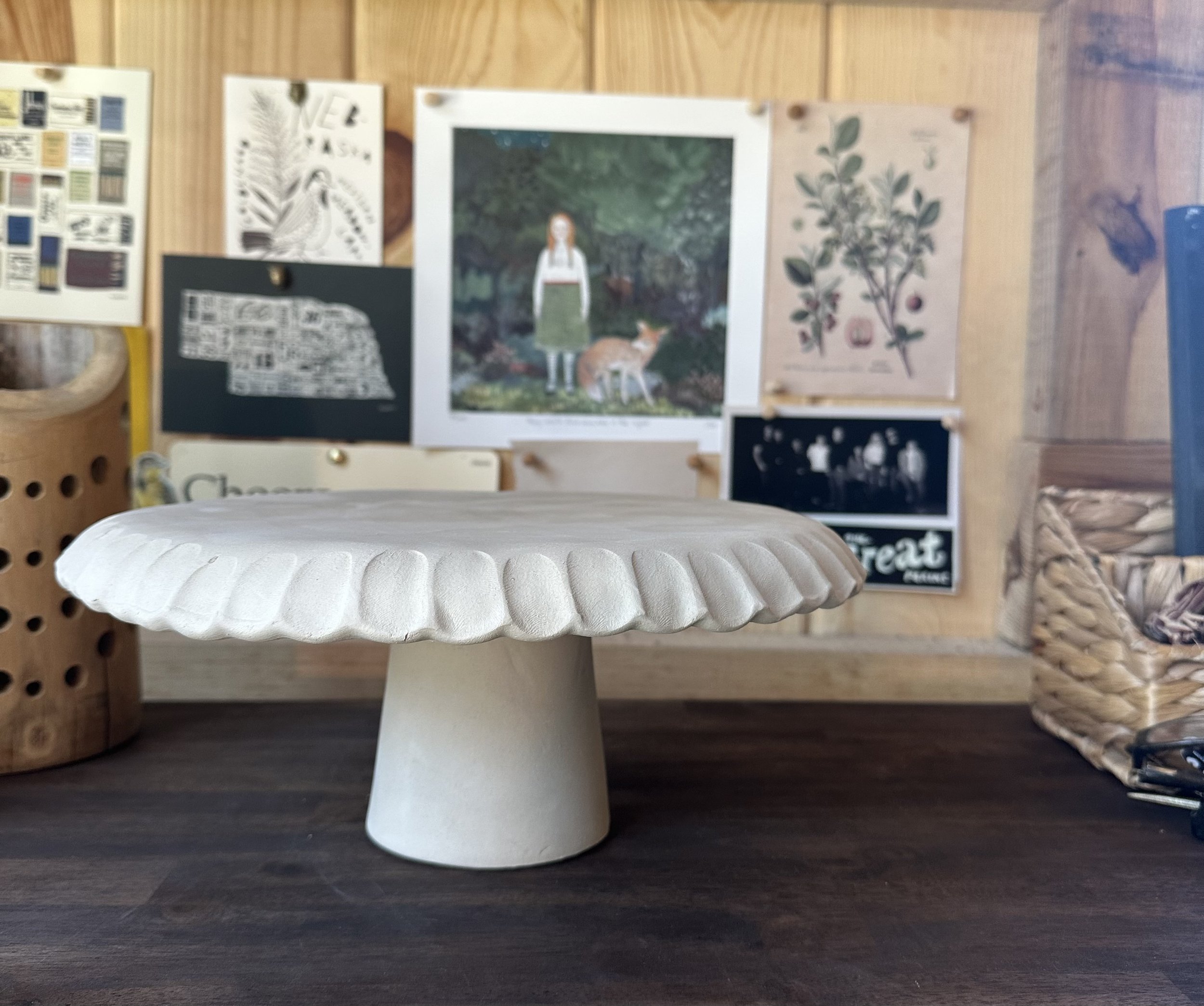 White decorative pedestal or cake stand with scalloped edge on a dark wooden surface, with various wall art and photographs in the background.