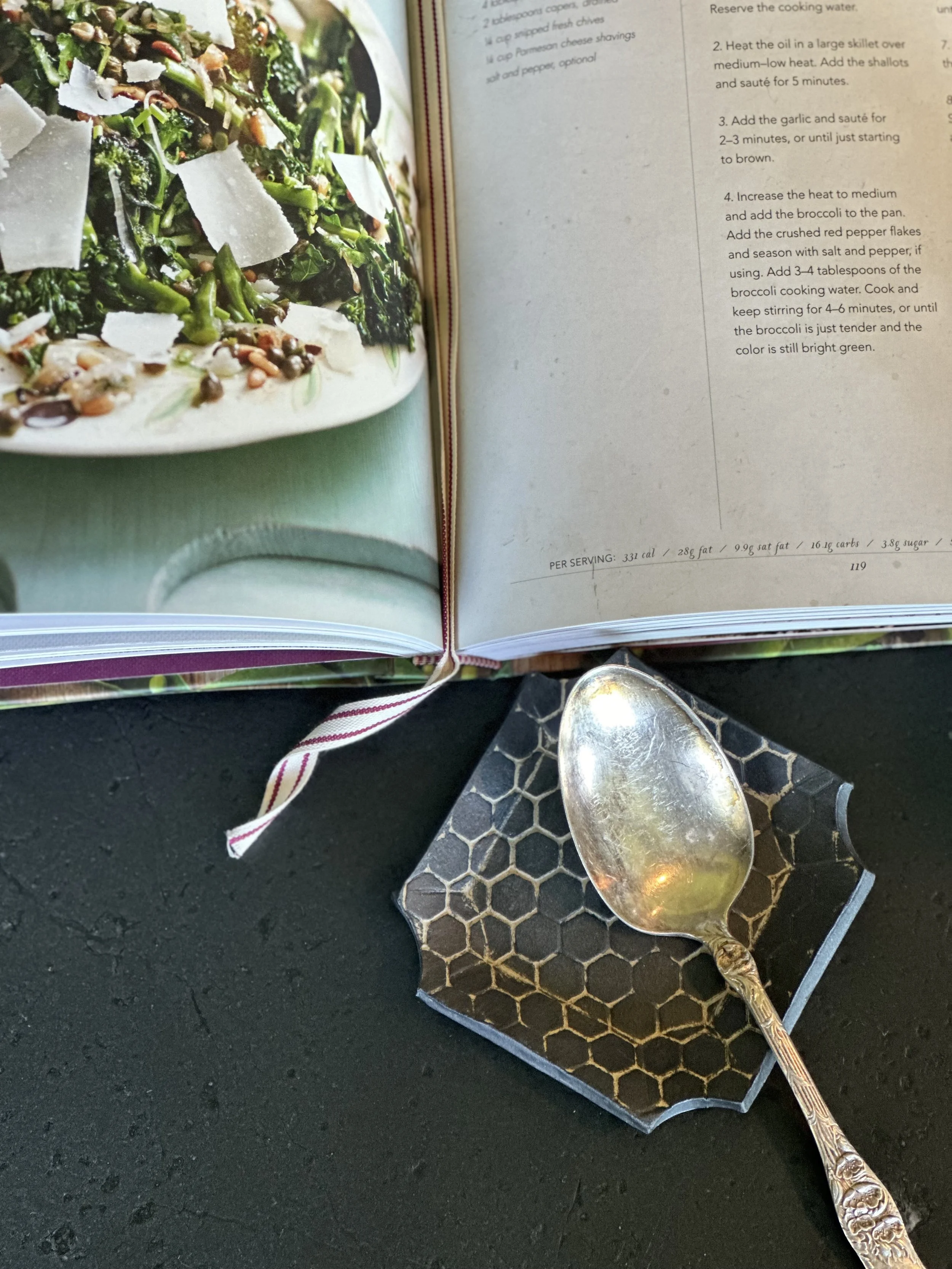 Open cookbook showing a photo of a green salad with shaved cheese and various toppings. A silver spoon rests on a black hexagon-patterned coaster on a black surface.